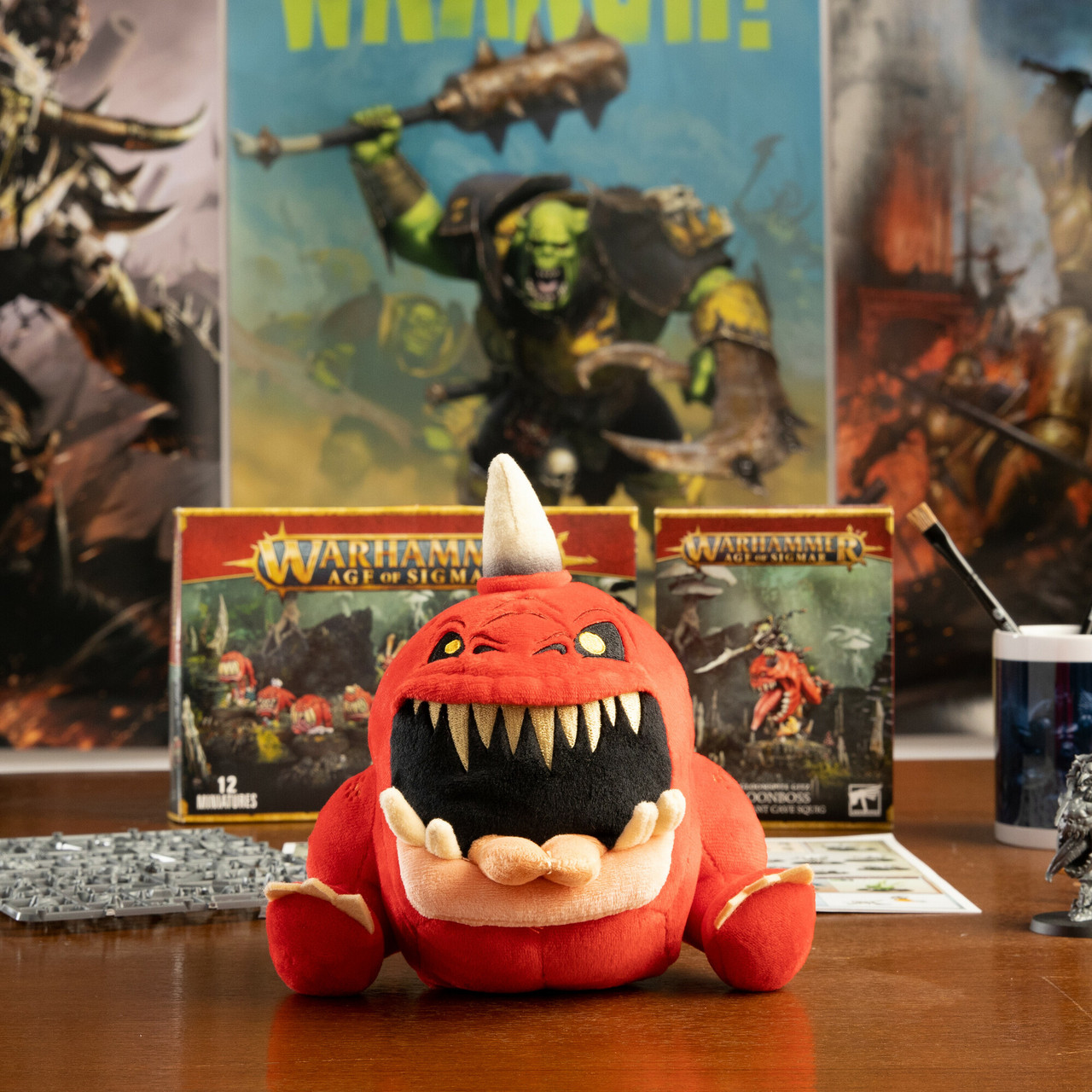 Une peluche rouge avec une grande bouche ouverte est devant deux boîtes Warhammer et une affiche. Des pinceaux et une figurine sont sur la table.
