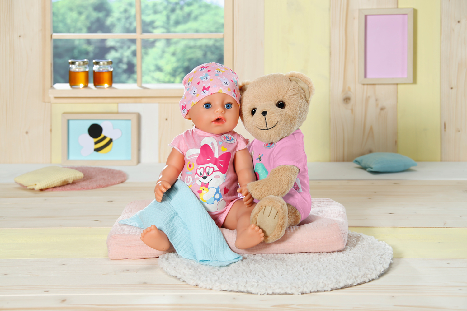 Eine Puppe und ein Teddybär sitzen auf einem rosa Kissen. Die Puppe trägt ein rosa Outfit und Hut. Im Hintergrund sind ein Fenster und ein Kunstrahmen.