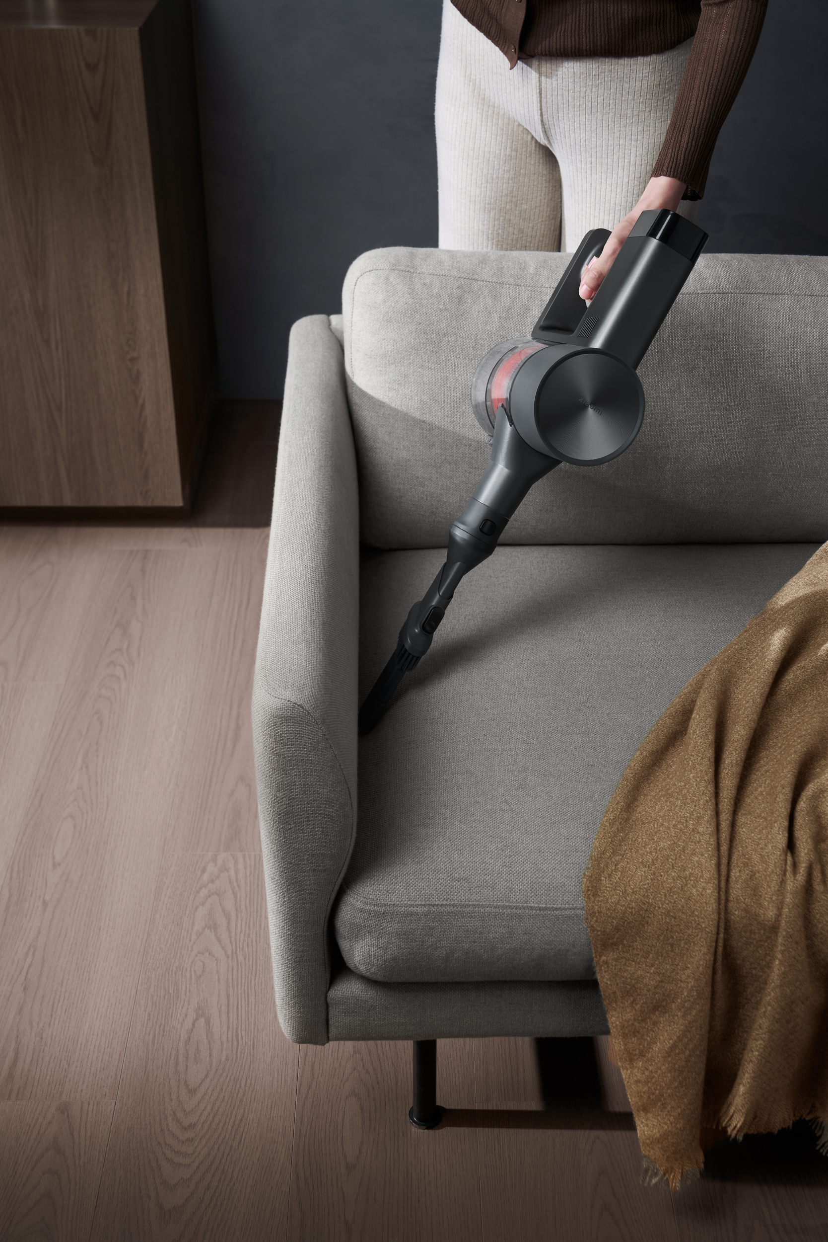Osoba używa szarego odkurzacza na szarej sofie. Na sofie leży brązowy koc, a podłoga jest drewniana.