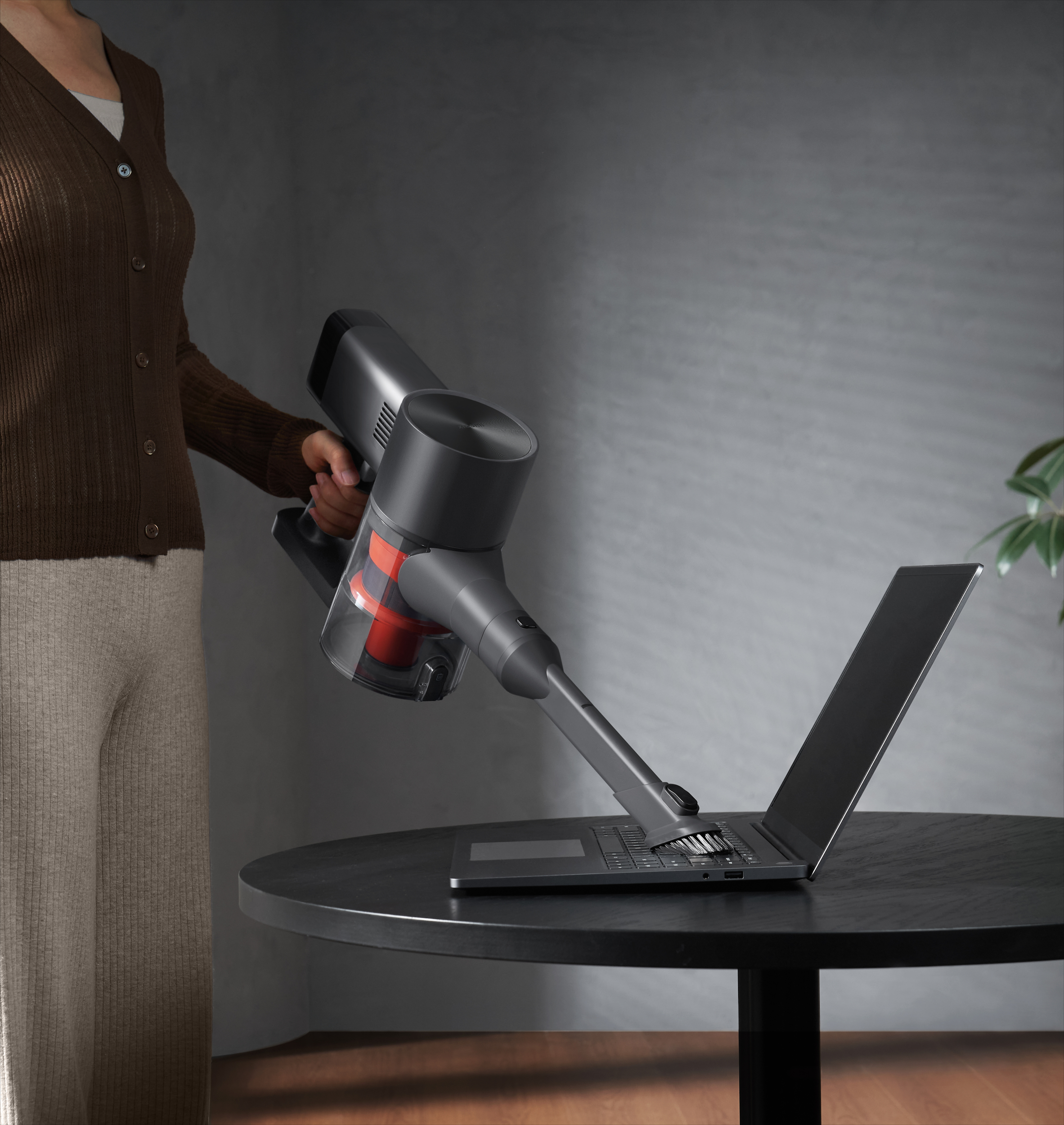 Osoba odkurza laptopa na czarnym stole szarym odkurzaczem. Tło to roślina i szara ściana.