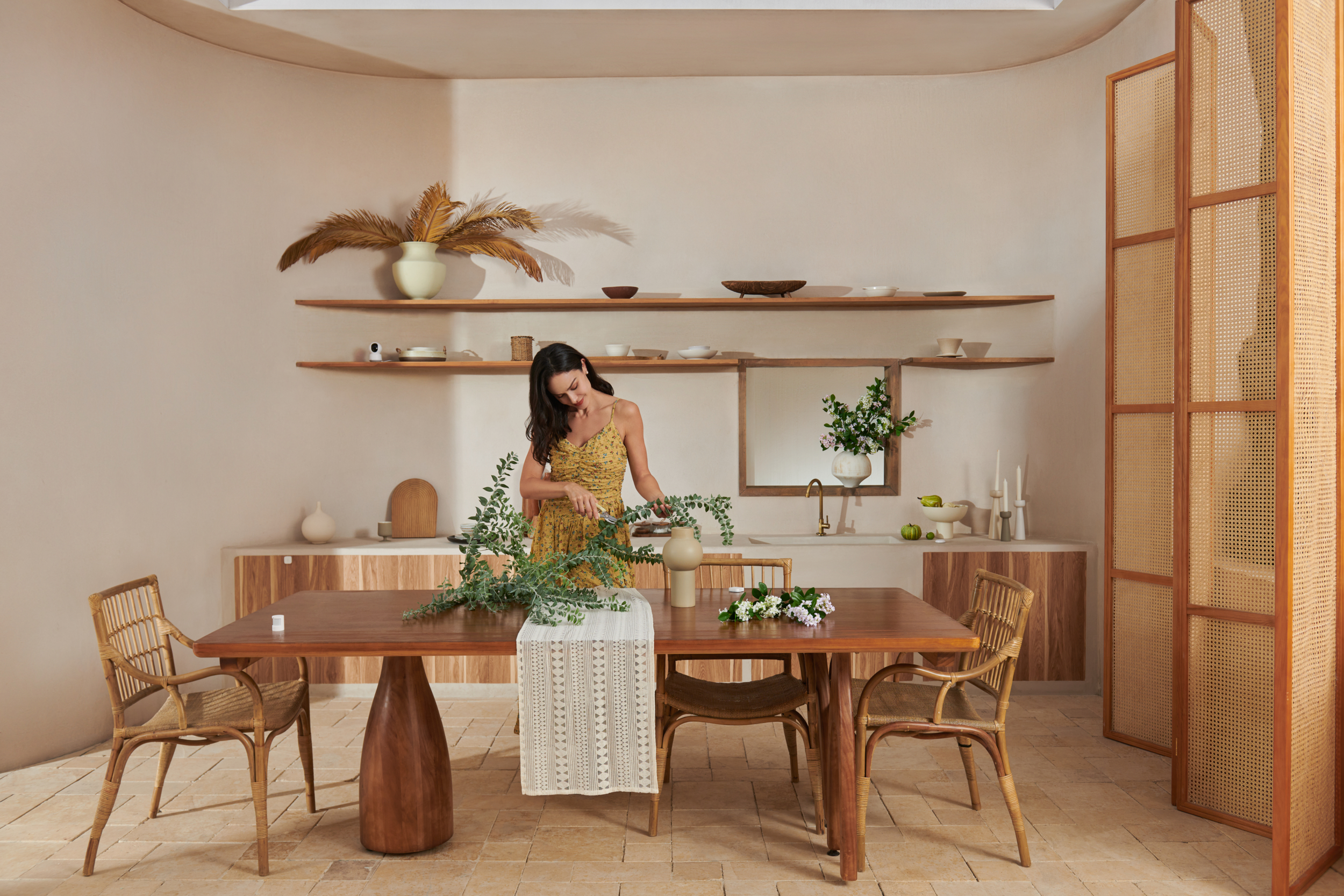 Een vrouw in een gele jurk schikt bloemen op een houten tafel. Een vaas en stoelen zijn in de buurt.