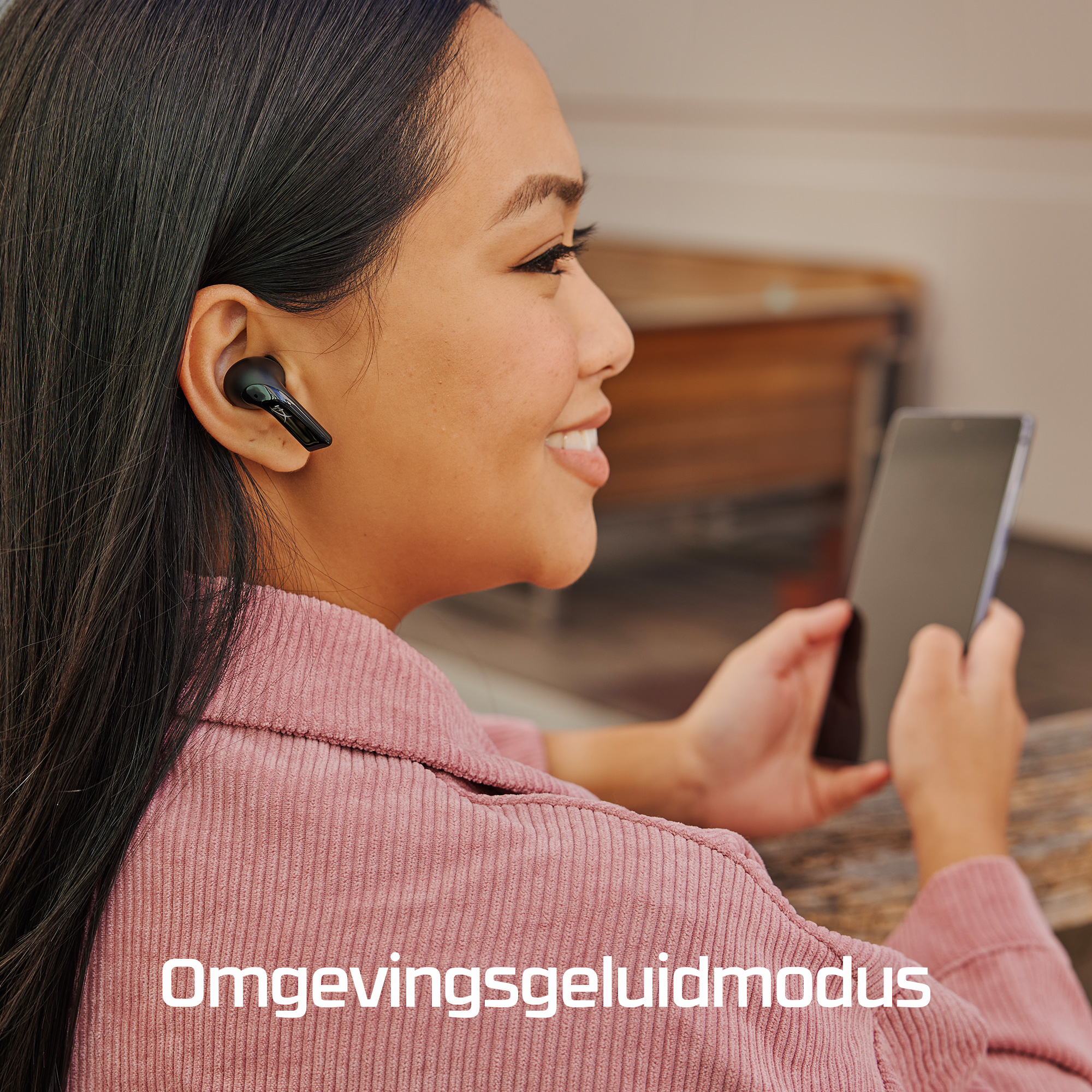 Kobieta ze słuchawkami uśmiecha się, trzymając telefon. Ma na sobie różową koszulę. Tekst 'Omgevingsgeluidmodus' jest na dole.