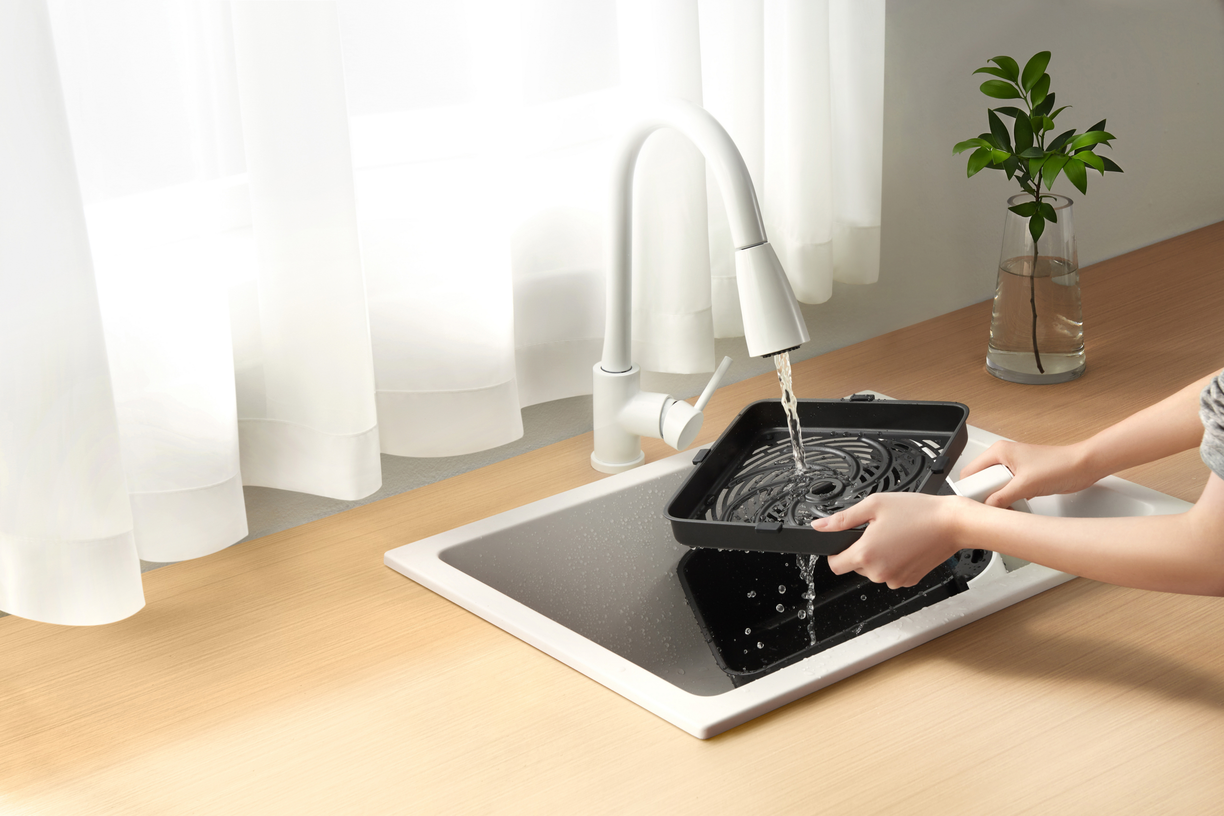 Osoba myje kosz do frytkownicy pod białym kranem. Roślina w wazonie po prawej. Białe zasłony.