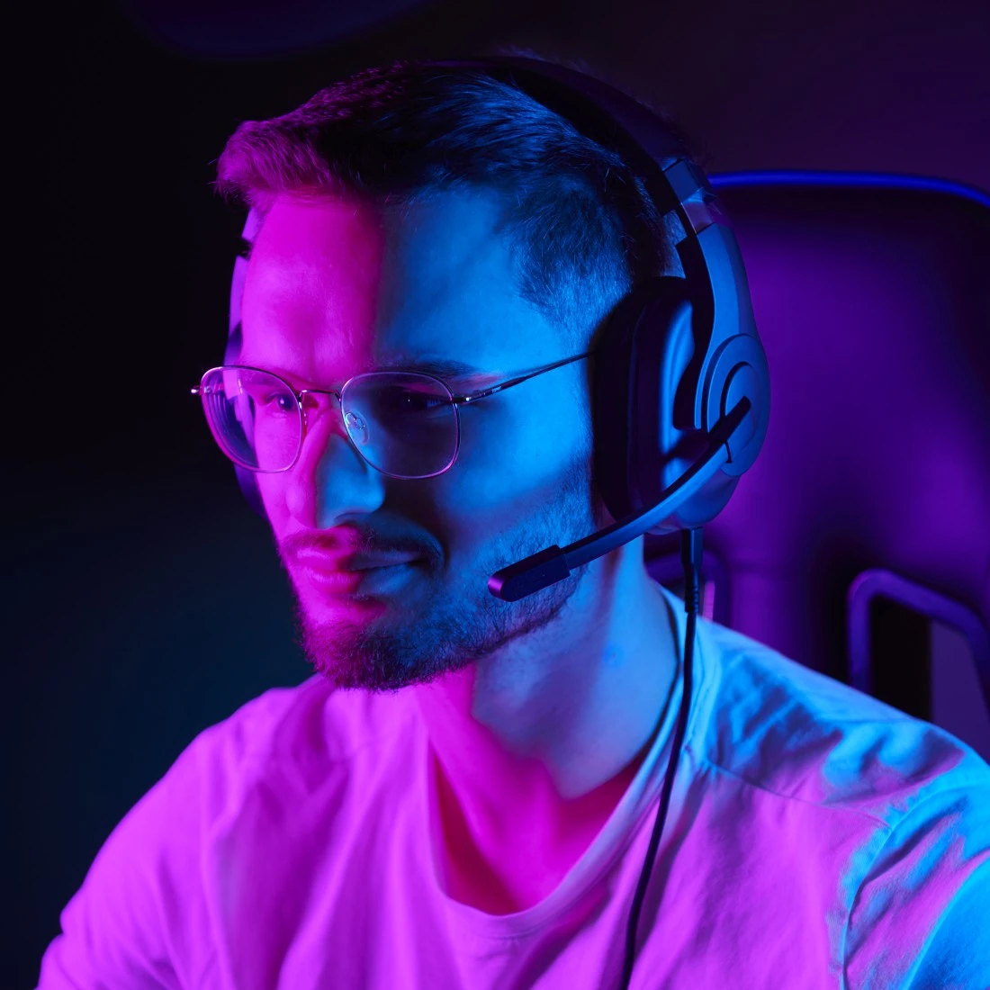 Un uomo con occhiali e cuffie, illuminato da luce blu e rosa. Indossa una camicia bianca ed è seduto.