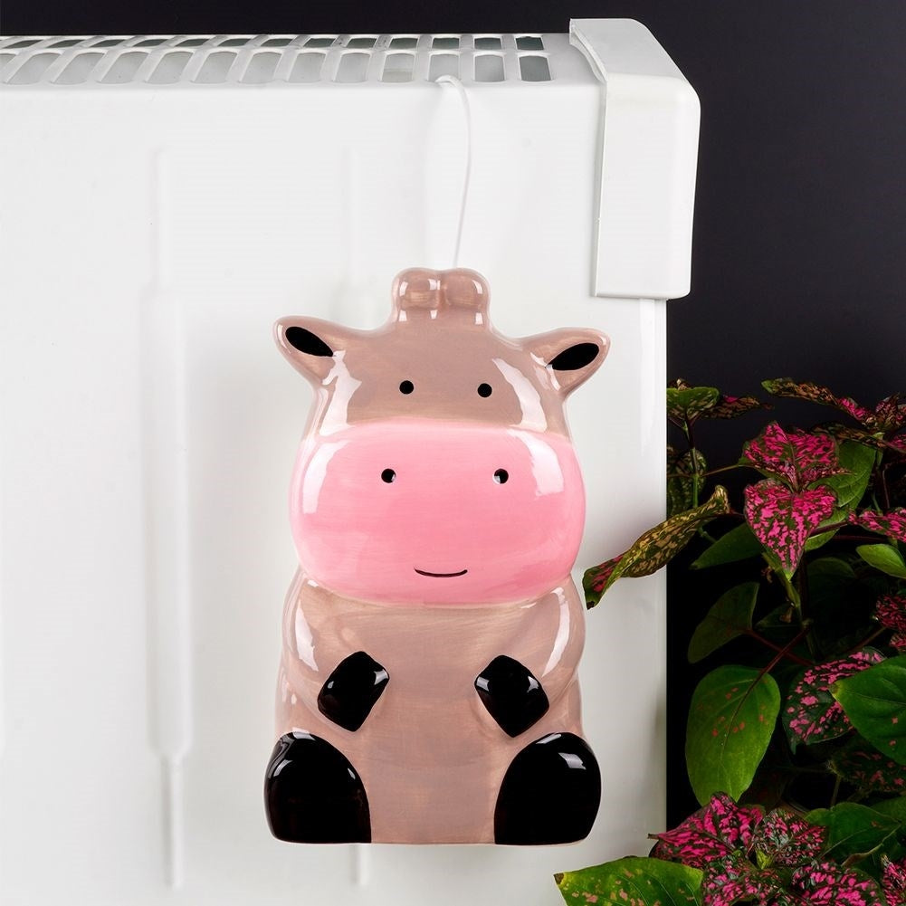 Un humidificador de cerámica con forma de vaca cuelga de un radiador blanco. La vaca es rosa y marrón claro, con detalles negros. Plantas rosas y verdes a la derecha.