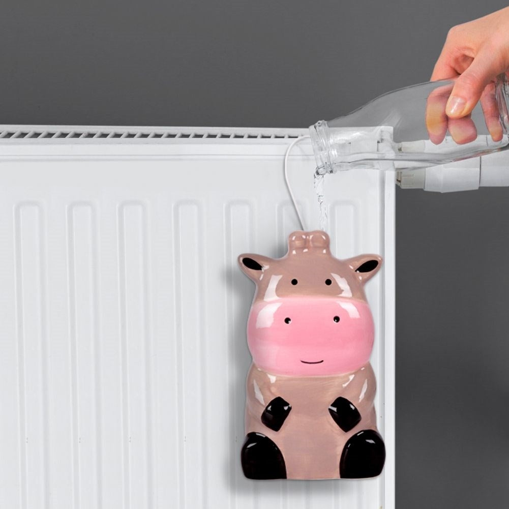 Un humidificador con forma de vaca está conectado a un radiador. Se vierte agua de una botella. El fondo es una pared gris.