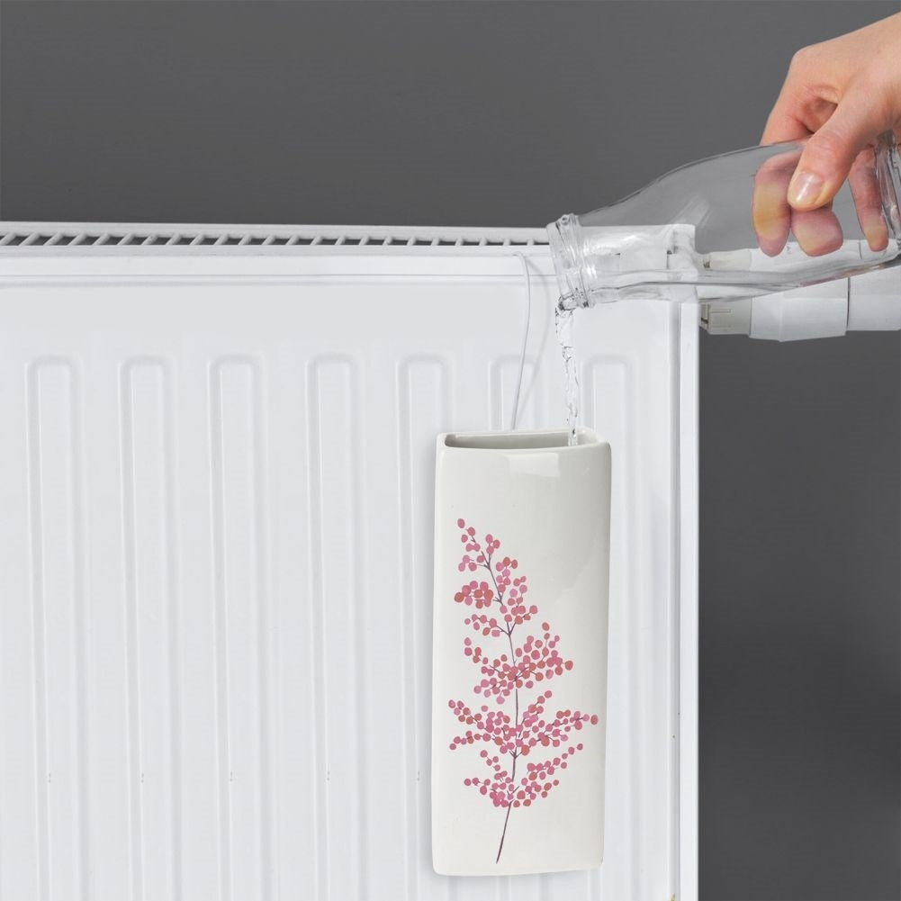 Una mano vierte agua de una botella en un jarrón blanco con diseño floral rosa, colgado en un radiador blanco.