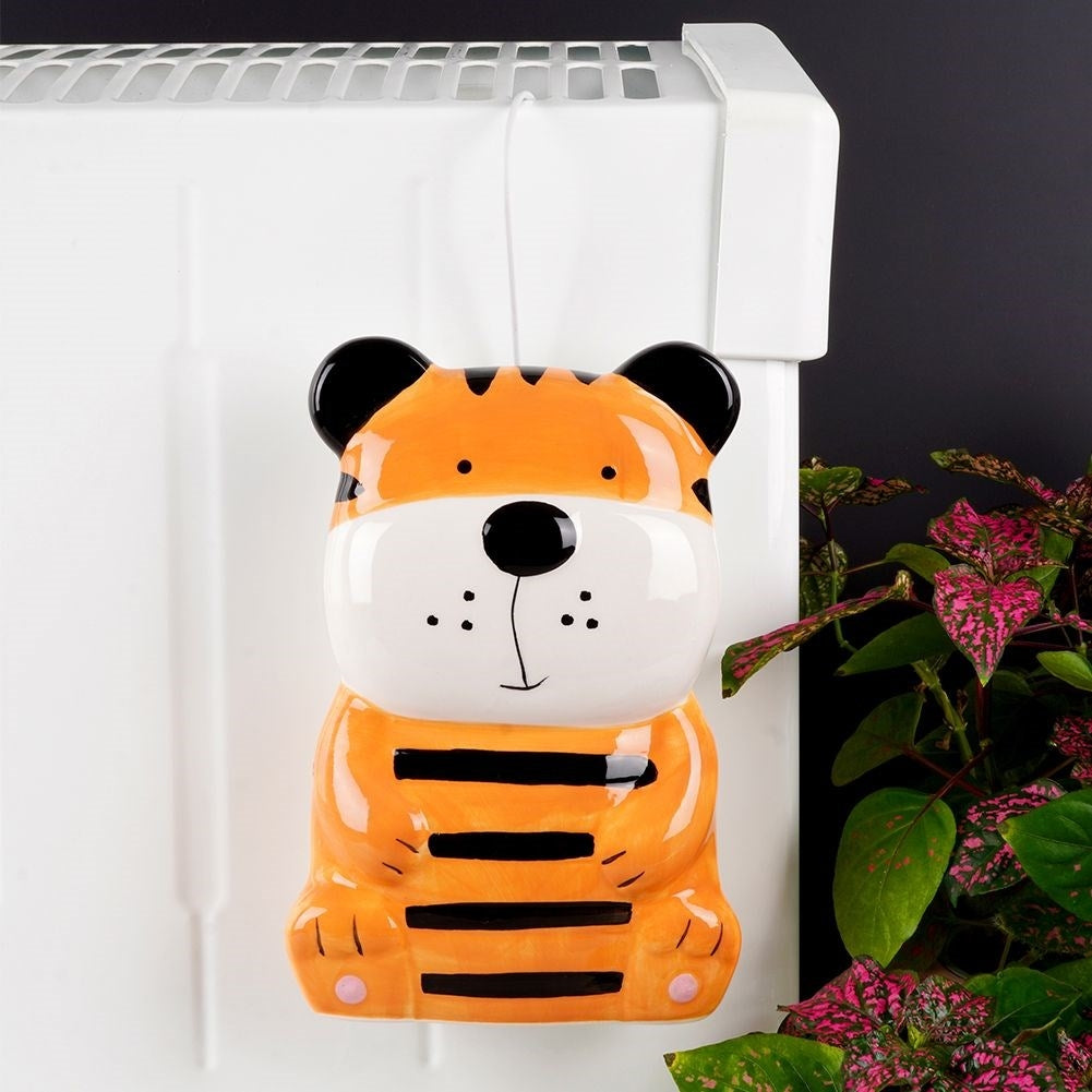Un adorno de tigre de cerámica cuelga de un radiador. Es naranja con rayas negras. Plantas con hojas rosadas a la derecha.