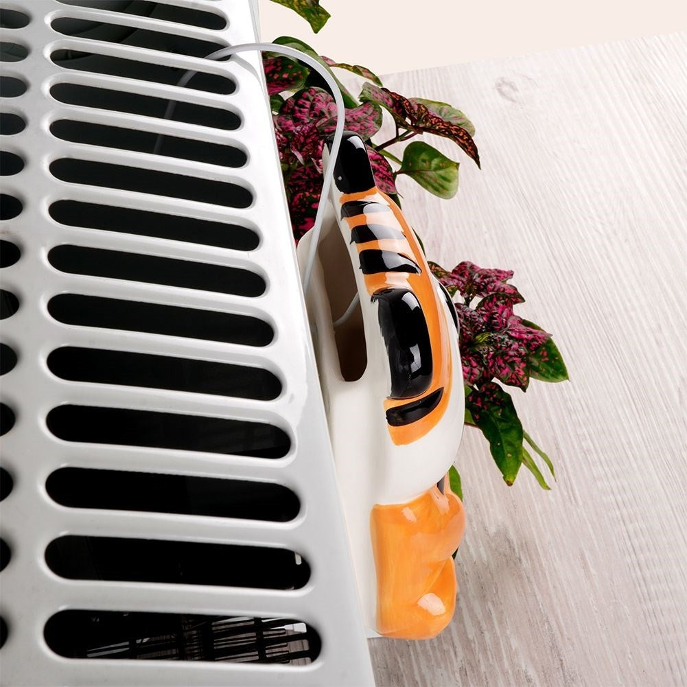 Una maceta decorativa con cara de tigre y una planta cuelga de un calentador blanco. El fondo es un panel de madera.
