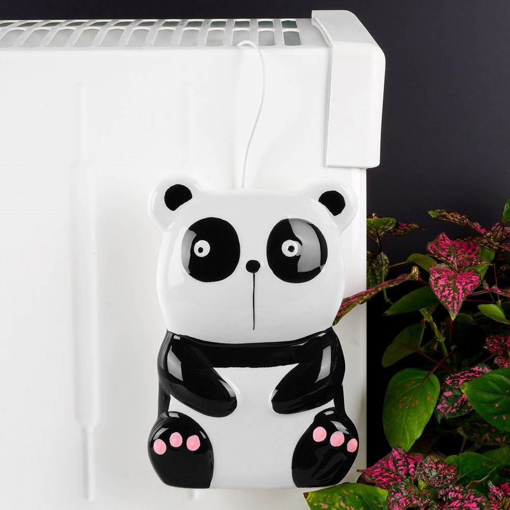 Una decoración con forma de panda cuelga de un calentador blanco. Tiene colores blanco y negro con detalles rosas. Hojas verdes y rosas a la derecha.