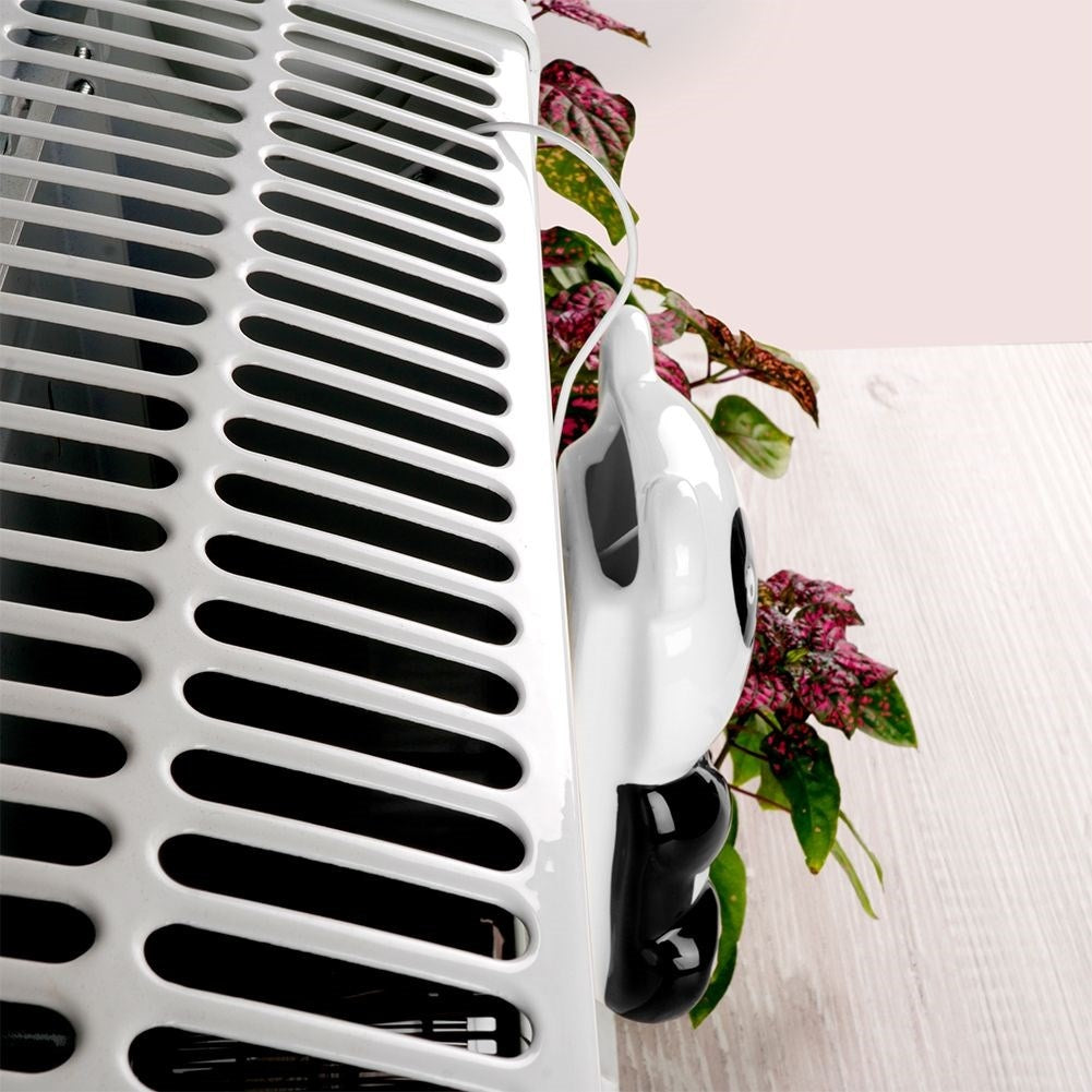 Una rejilla de ventilación blanca con ranuras negras. Una pieza decorativa blanca y negra cuelga cerca de una planta con hojas rosas y verdes.