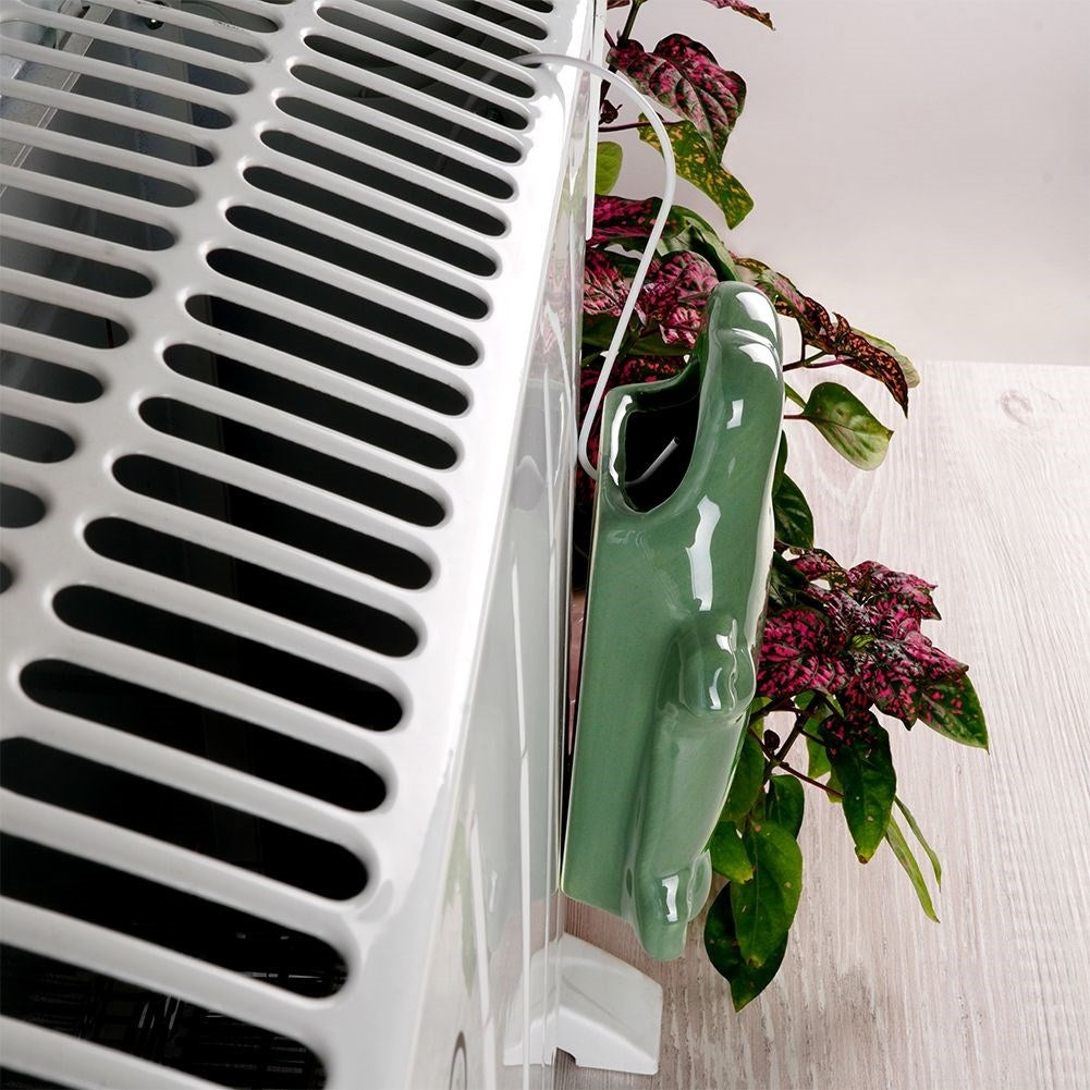 Un radiador blanco con un macetero de cerámica verde. La planta tiene hojas rojas y verdes. El fondo es una pared blanca.