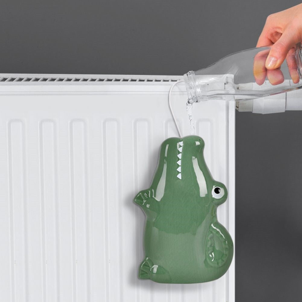 Un humidificador verde con forma de cocodrilo cuelga de un radiador. Se vierte agua en él desde una botella de vidrio. Fondo gris.
