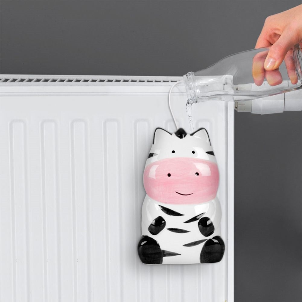 Un humidificador con forma de cebra cuelga de un radiador, se vierte agua de una botella. La cebra tiene hocico rosa, rayas negras y patas.