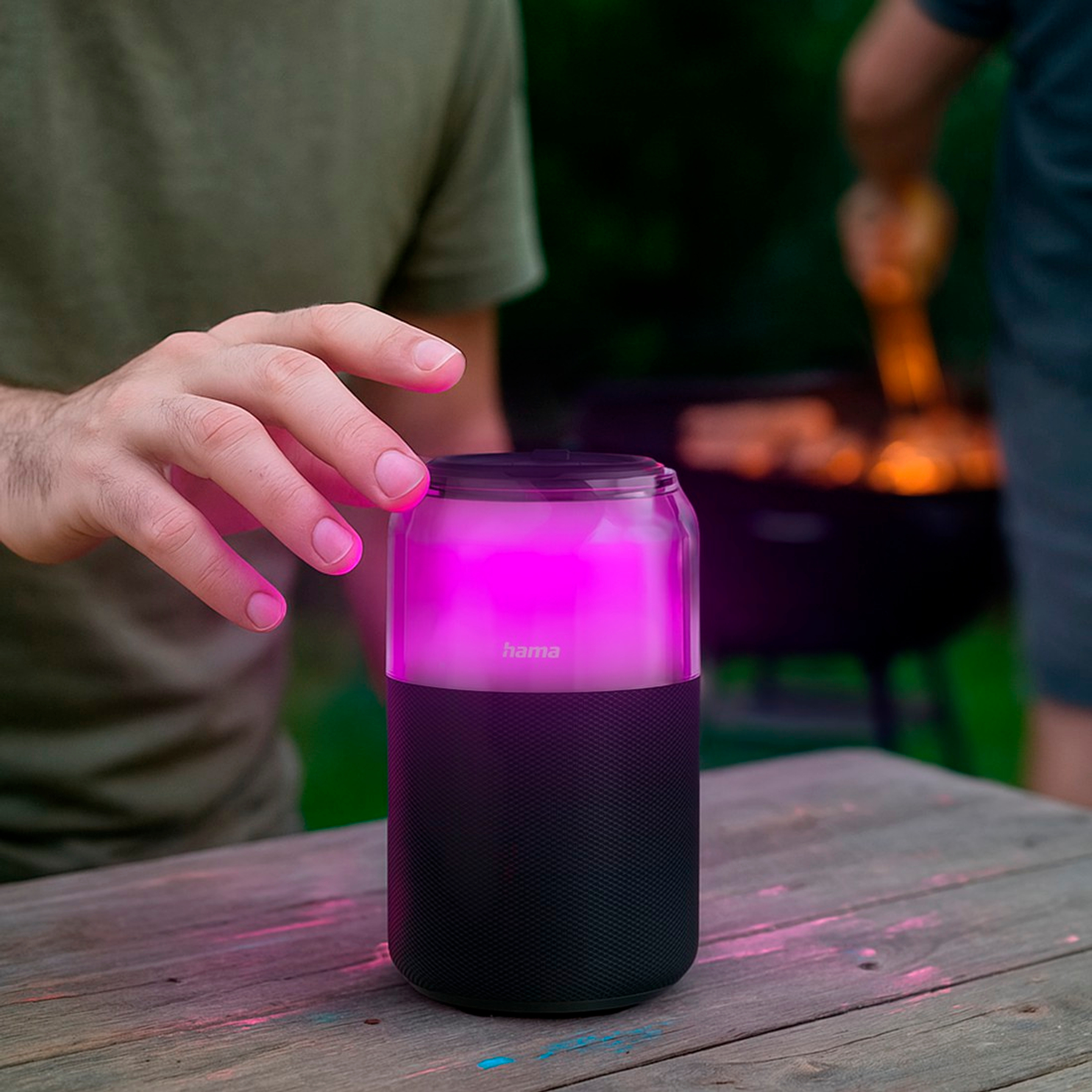 La mano di una persona tocca un altoparlante rosa brillante su un tavolo di legno. Uno schermo e un'altra persona sono sfocati sullo sfondo.