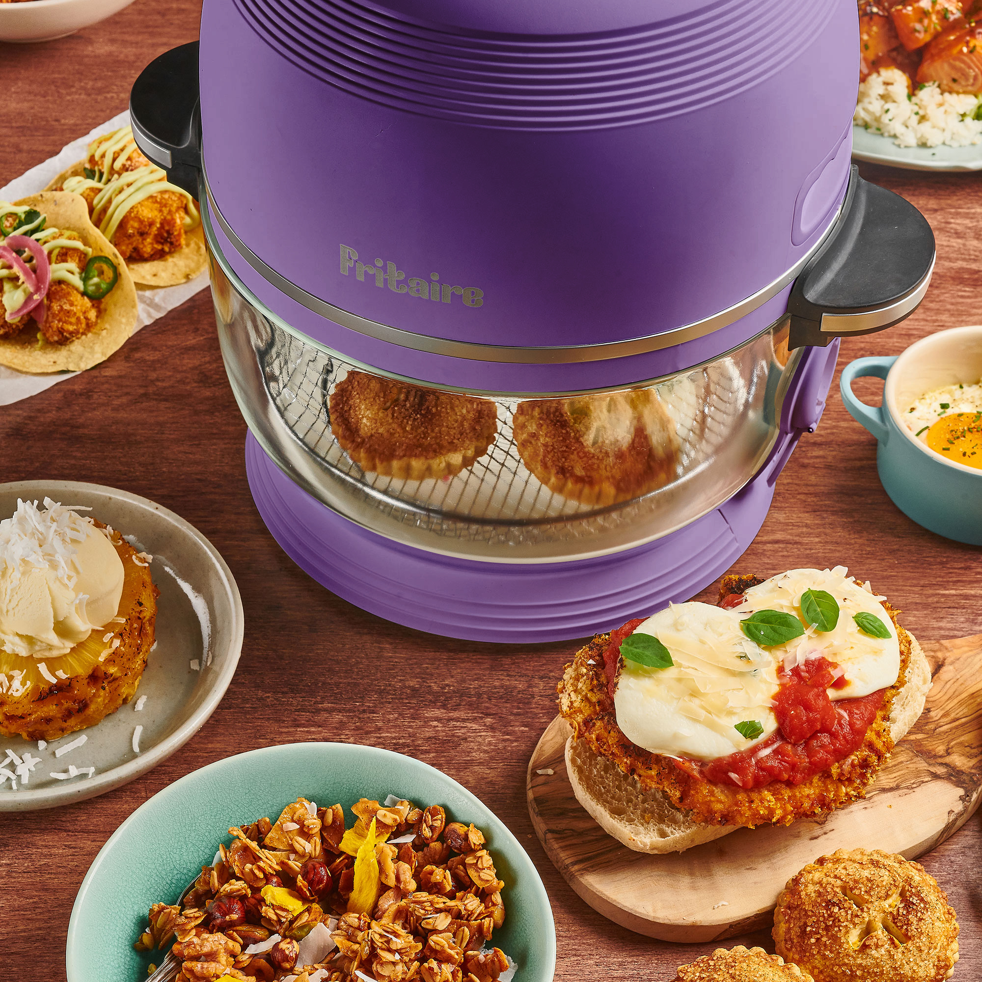 Ein lila Airfryer mit Speisen und verschiedenen Gerichten wie Tacos, Müsli und einem Caprese-Sandwich auf einem Holztisch.