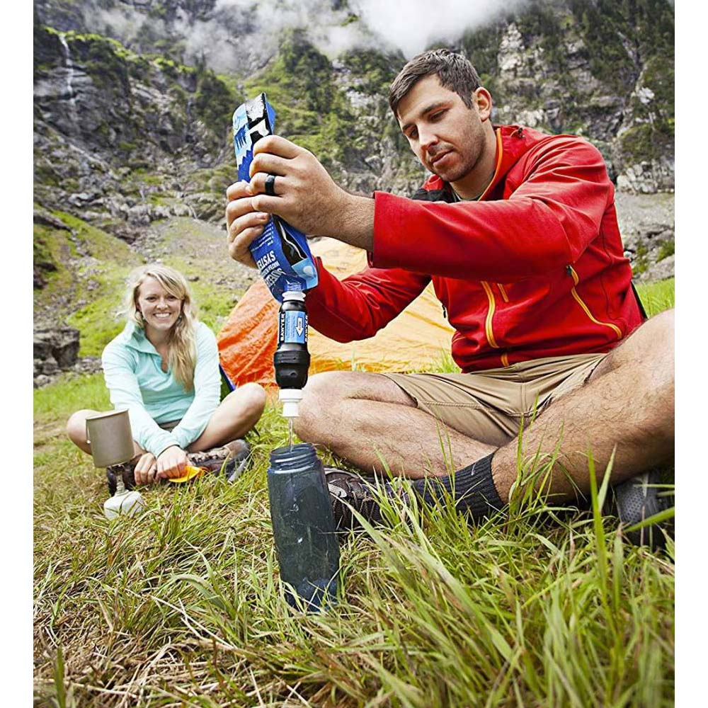 Uomo in giacca rossa filtra acqua in una bottiglia. Donna sorride dietro. Scena di campeggio con erba e montagne.