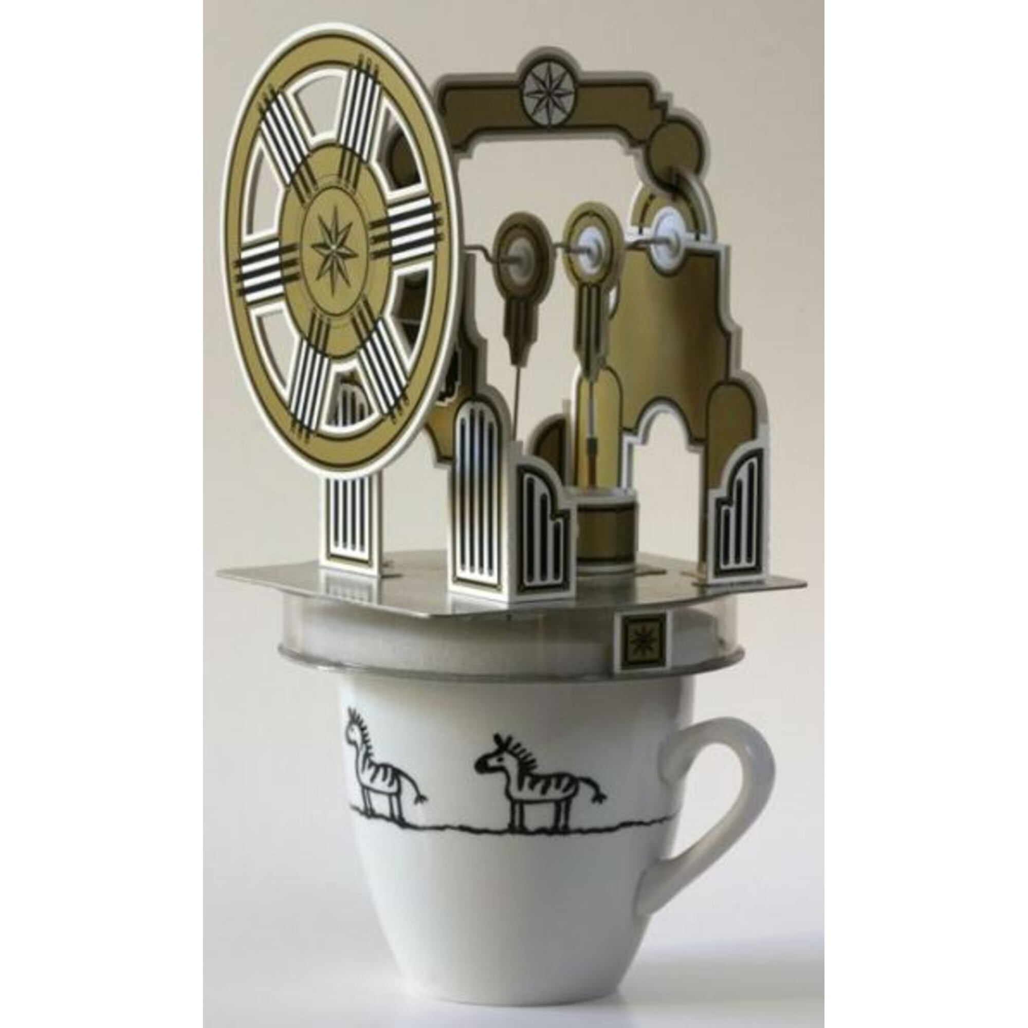 Ein dekoratives Objekt steht auf einer Tasse. Das Objekt ist gold-silberfarben und hat ein Raddesign. Die Tasse hat Zebrazeichnungen.