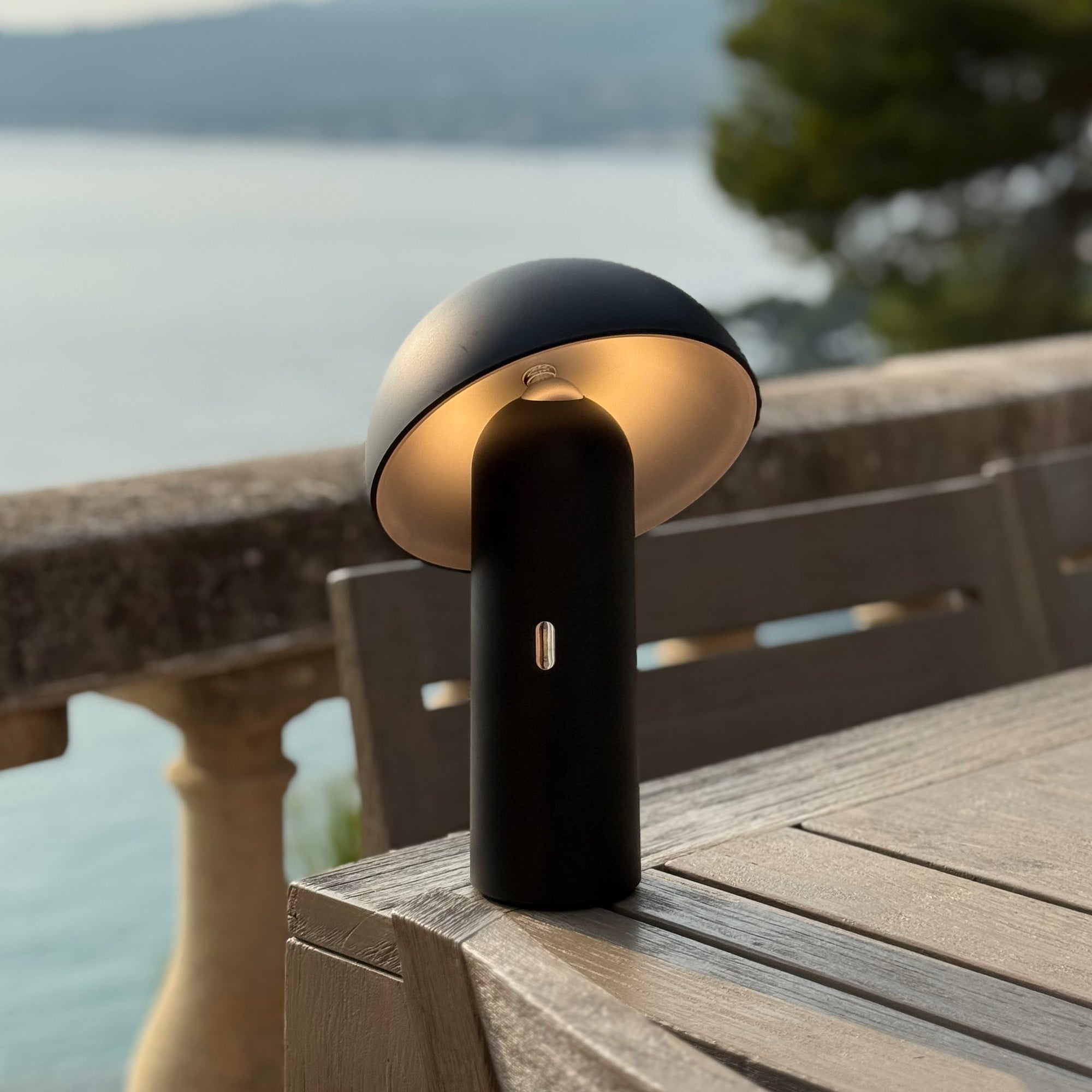 Eine schwarze pilzförmige Lampe steht auf einem Holztisch, im Hintergrund sind Meer und Bäume zu sehen.