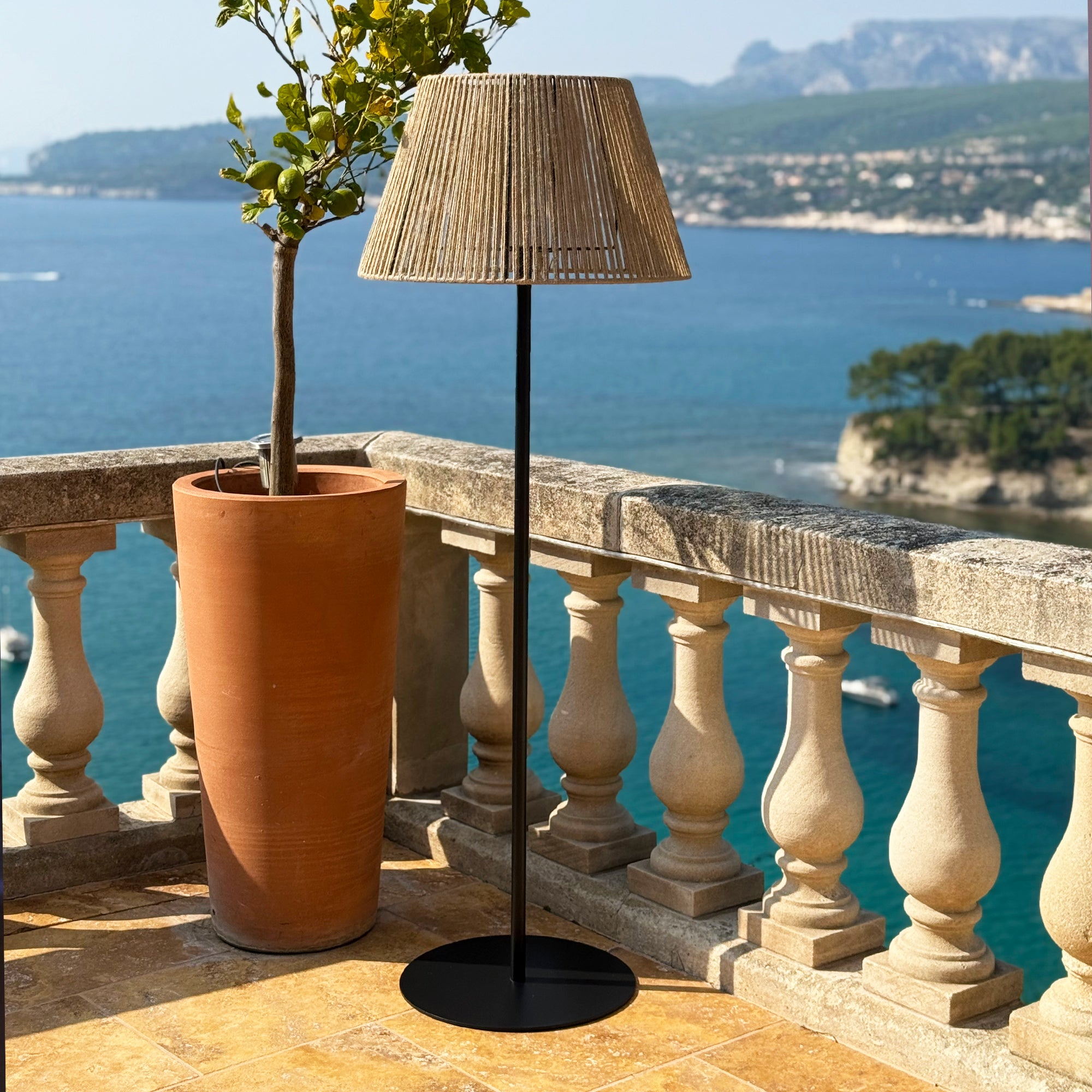 Eine Aussicht am Meer mit einem Baum im Topf, einer Lampe und einer Steinbalustrade. Das Meer ist blau und der Himmel klar.