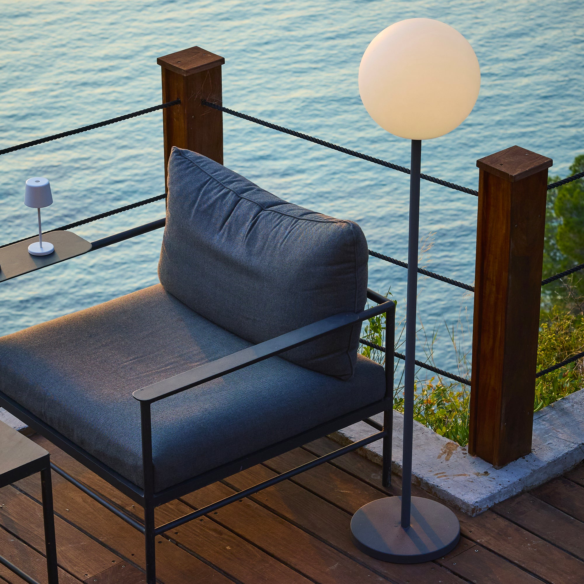 Scena all'aperto con sedia grigia, lampada e tavolo. Mare e decking in legno visibili. Luce serale.