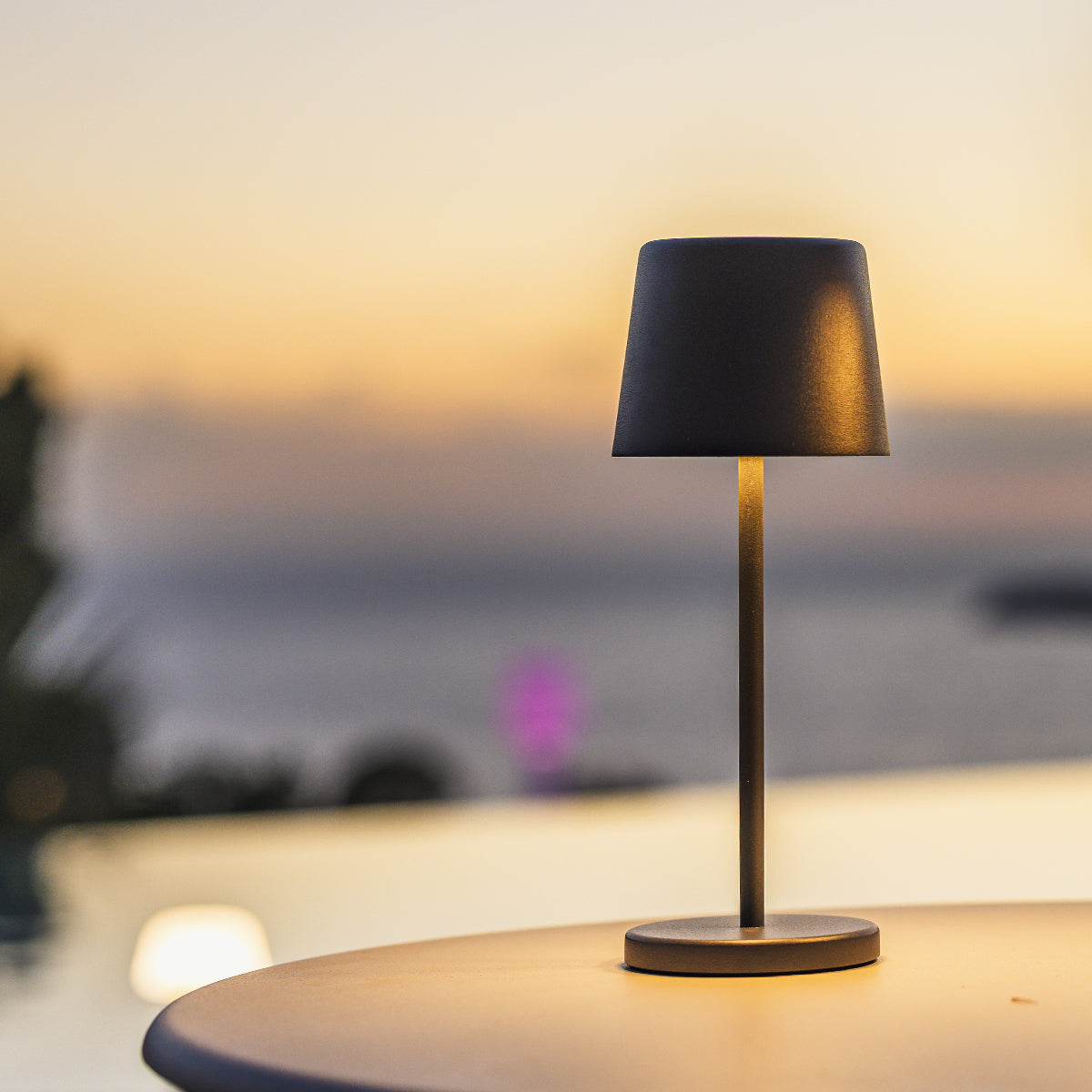 Eine schwarze Lampe mit Lampenschirm steht auf einem Tisch. Im Hintergrund sind Wasser und ein Sonnenuntergang zu sehen.