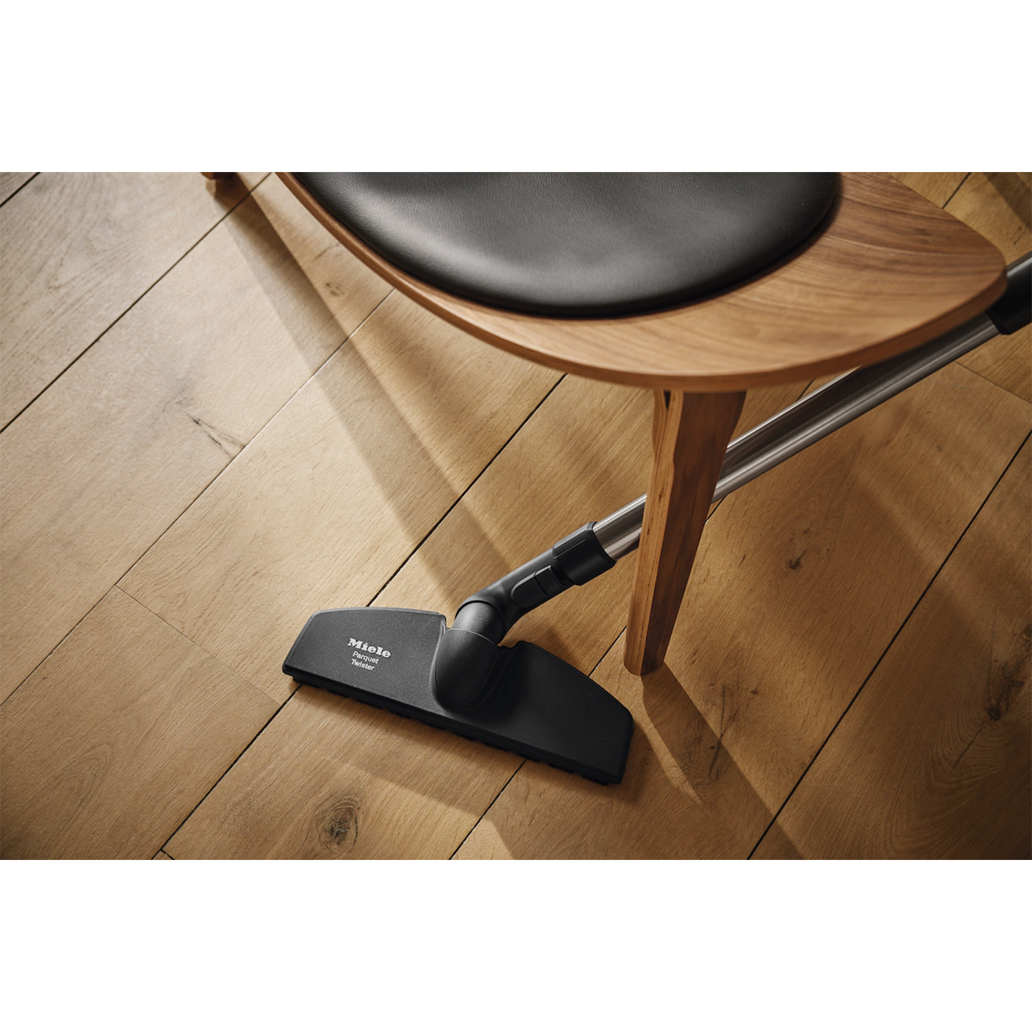 Głowica odkurzacza znajduje się na drewnianej podłodze w pobliżu drewnianego krzesła. Podłoga jest jasnobrązowa. Krzesło ma czarne siedzisko.