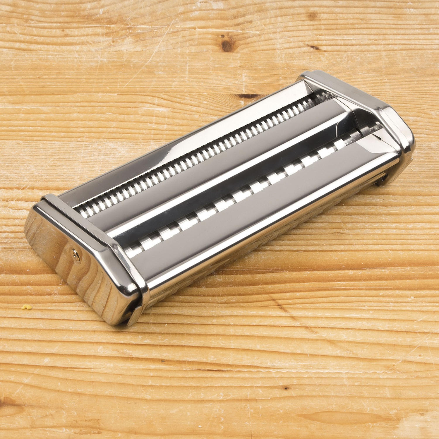 Una macchina per la pasta in metallo su una superficie di legno. La macchina è argentata e ha rulli. Il legno ha venature visibili.
