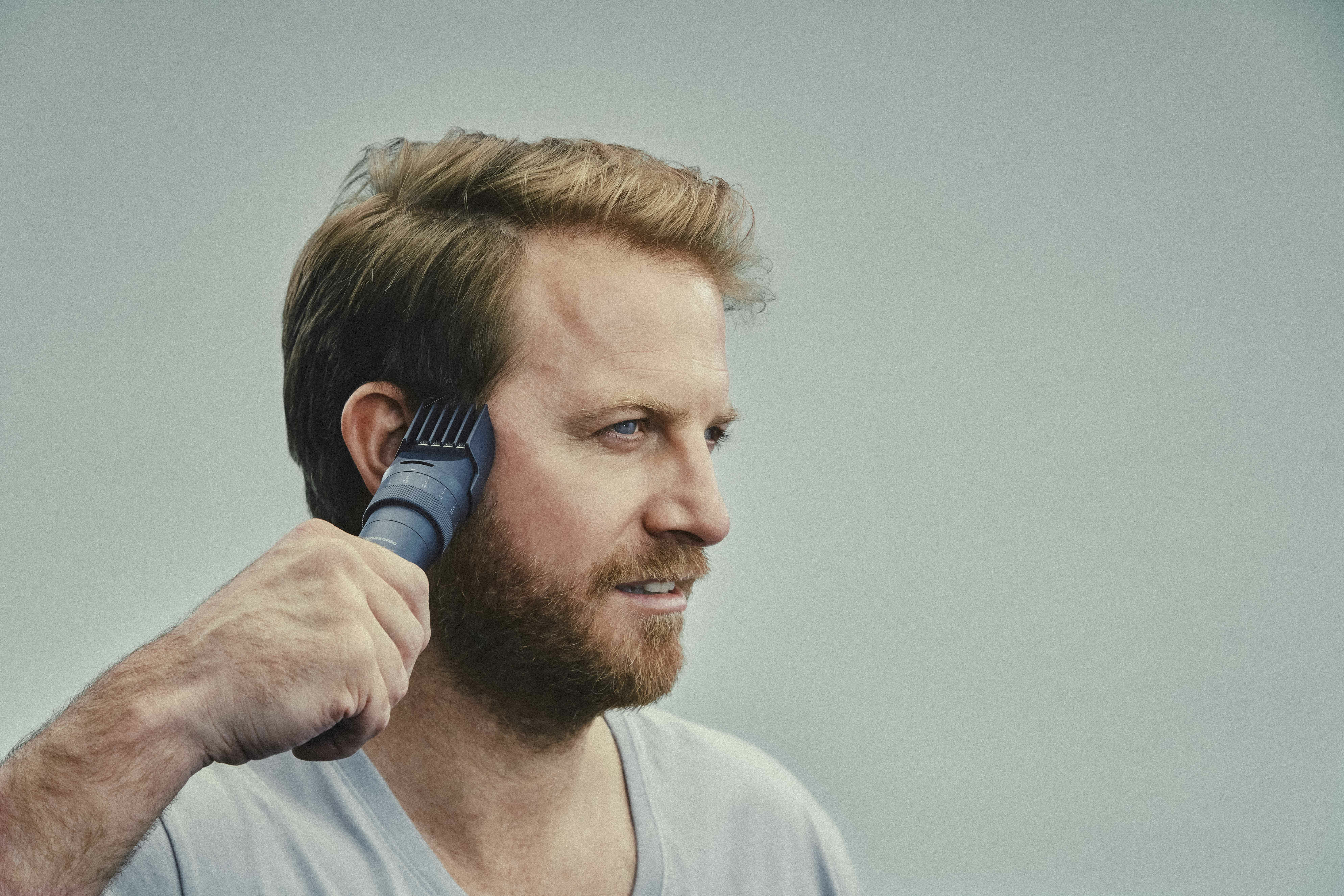 Ein Mann rasiert sich mit einem elektrischen Trimmer die Haare. Er hat rotes Haar, einen Bart und blaue Augen.