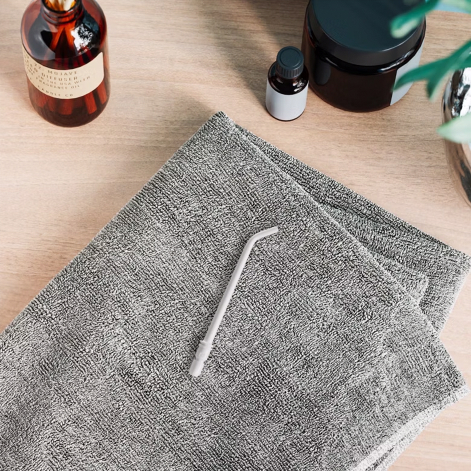 Graues Handtuch mit grauer Düse, braune Diffusorflasche, kleine Flasche und ein schwarzes Glas auf Holzoberfläche.