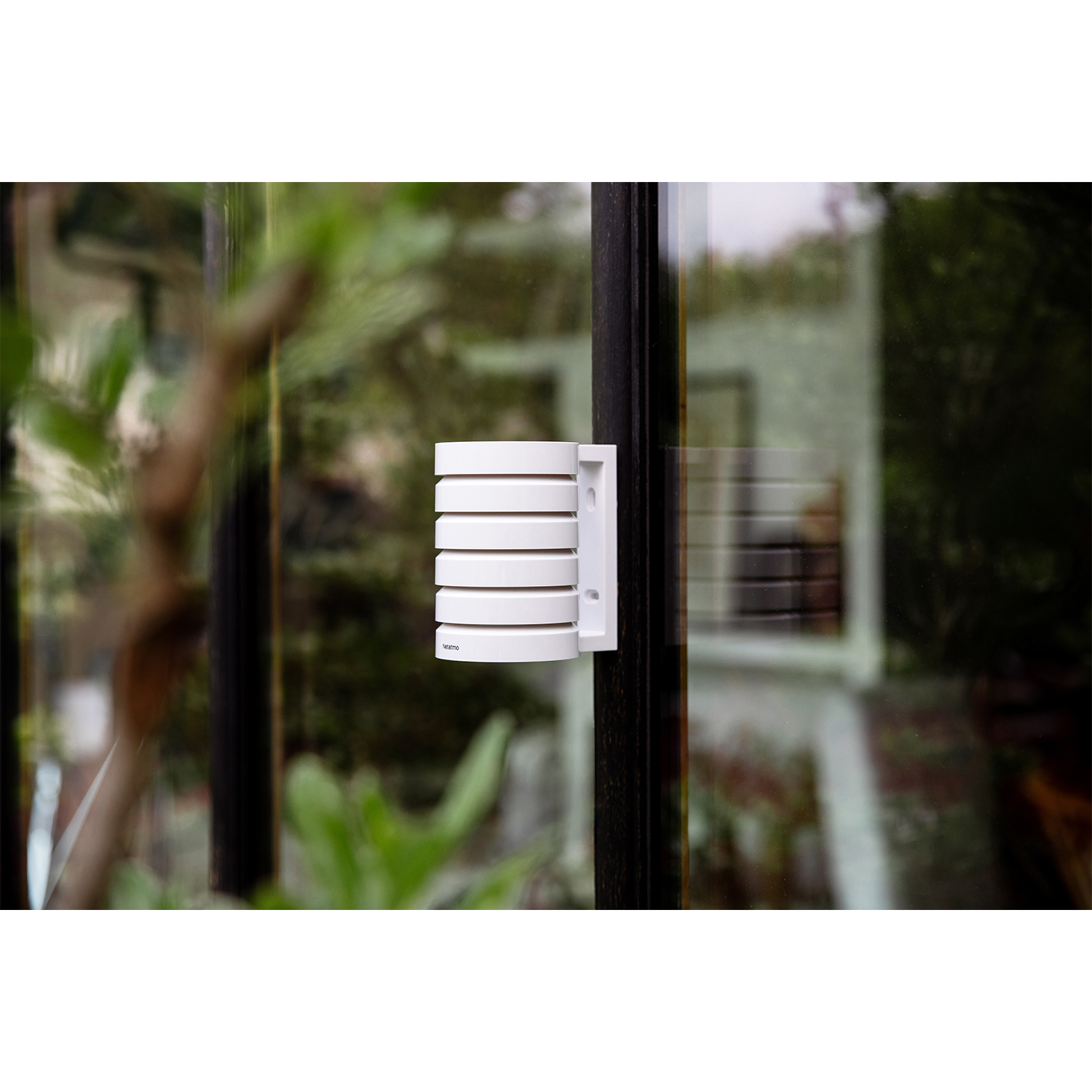 A white, cylindrical weather station is mounted on a dark window frame. The background shows blurred green foliage.
