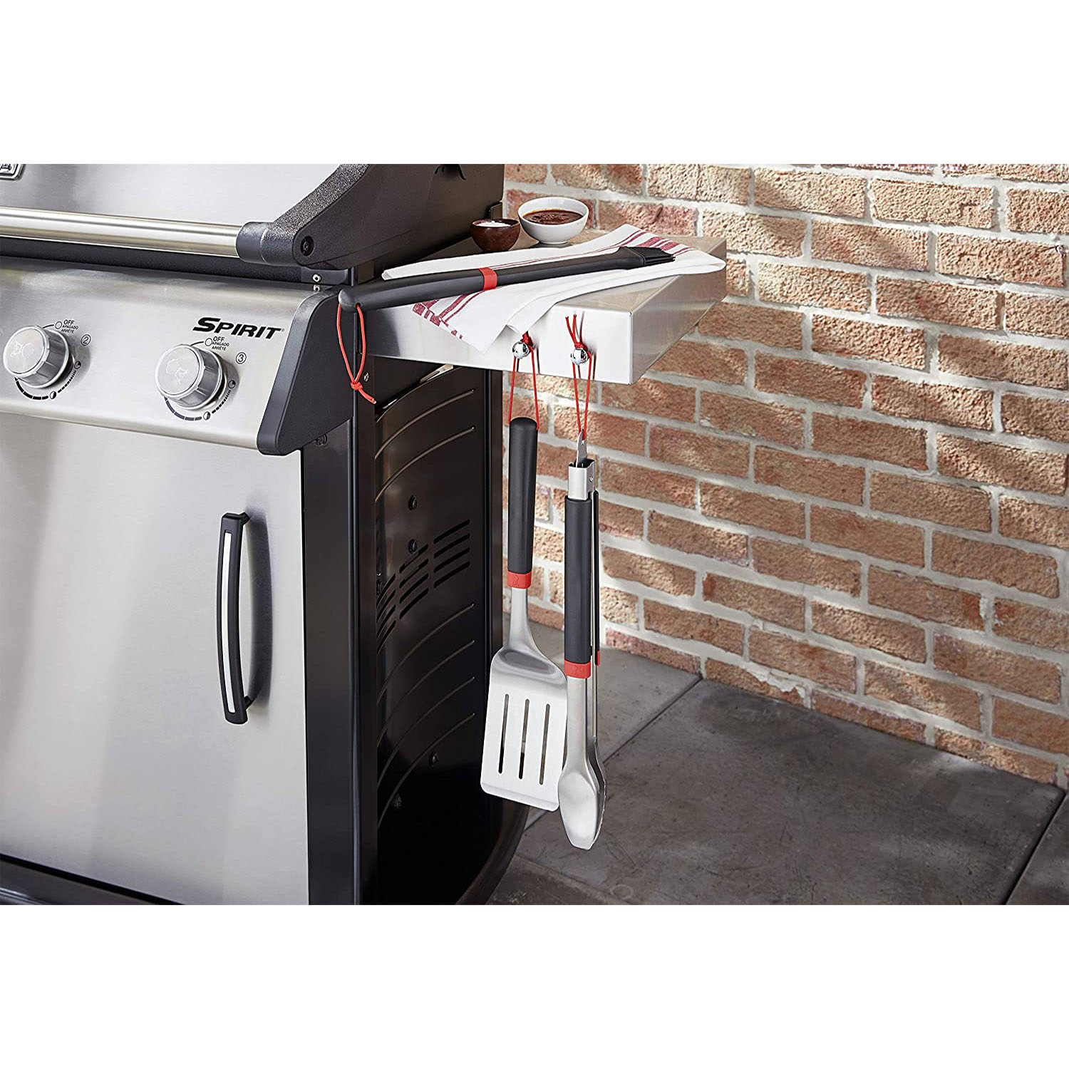Grill gazowy ze stali nierdzewnej z wiszącymi narzędziami. W tle ceglana ściana. Wiszą szczypce, szpatułka i pędzel.