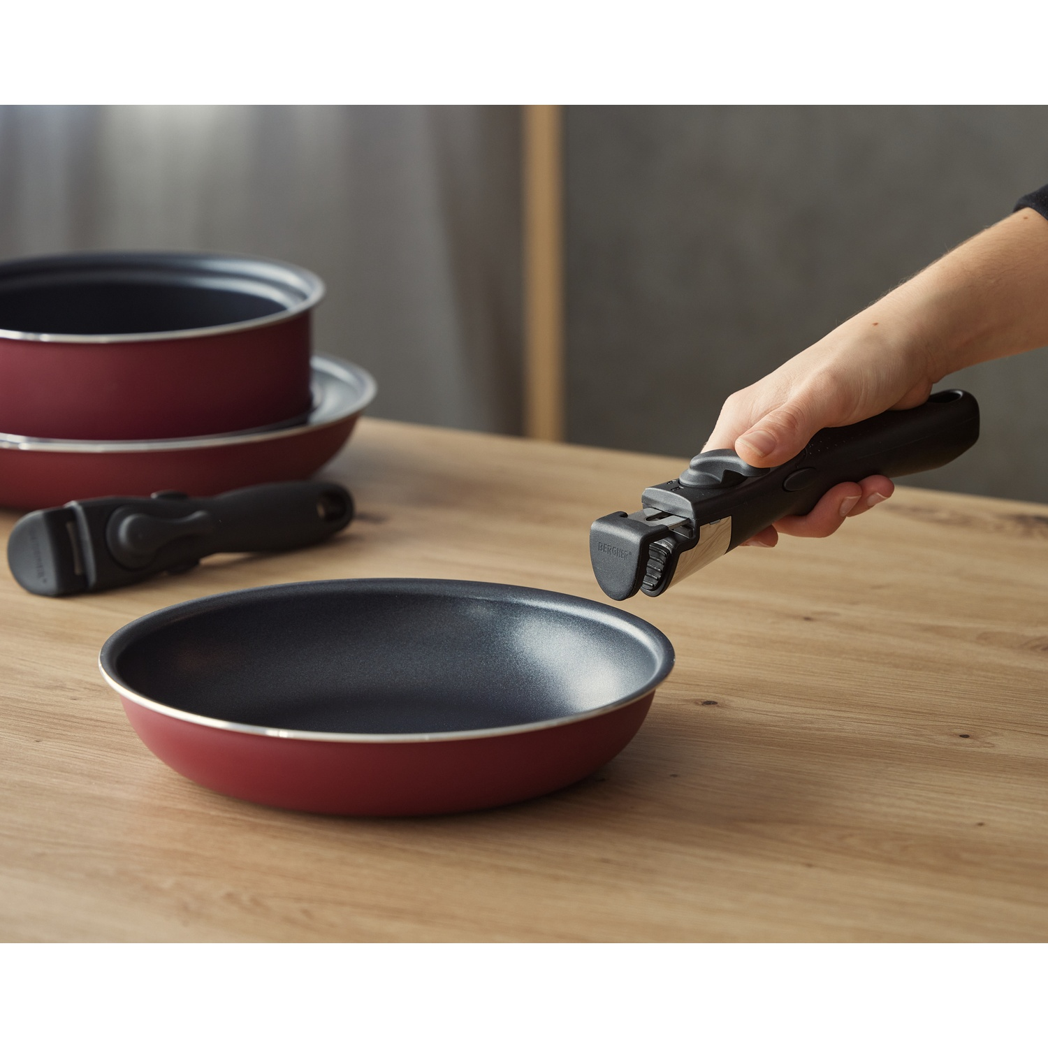 Una mano conecta un mango negro a una sartén roja sobre una mesa de madera. Se ven otras sartenes y ollas en el fondo.