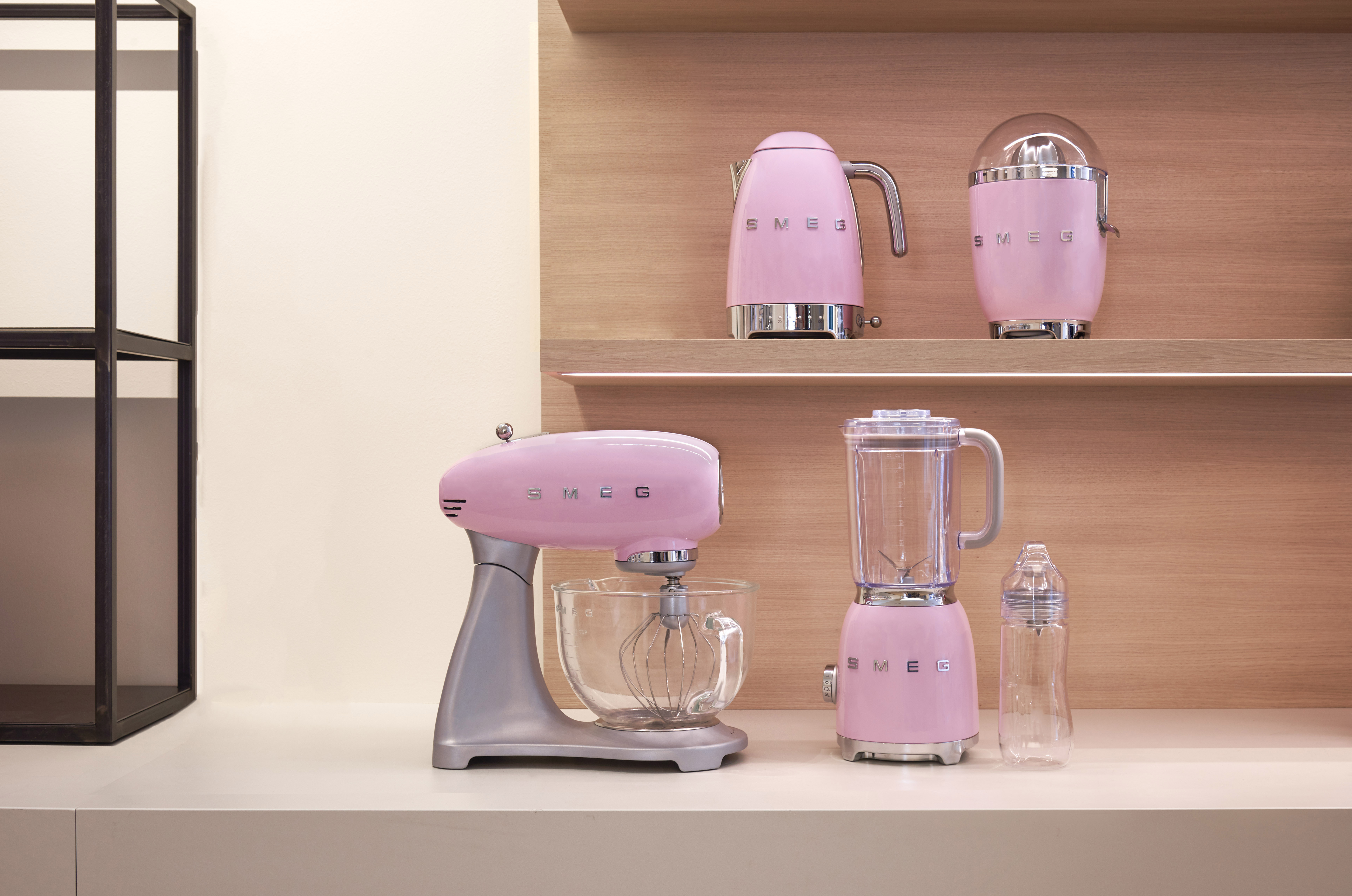 Pembe SMEG mutfak aletleri: mikser, blender, su ısıtıcısı ve mutfak robotu. Ahşap raflar ve beyaz tezgah.