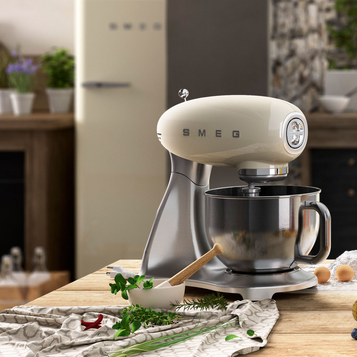 Un mixer da cucina SMEG color crema è su un tavolo di legno con erbe, un mortaio e uova. Un frigorifero sullo sfondo.
