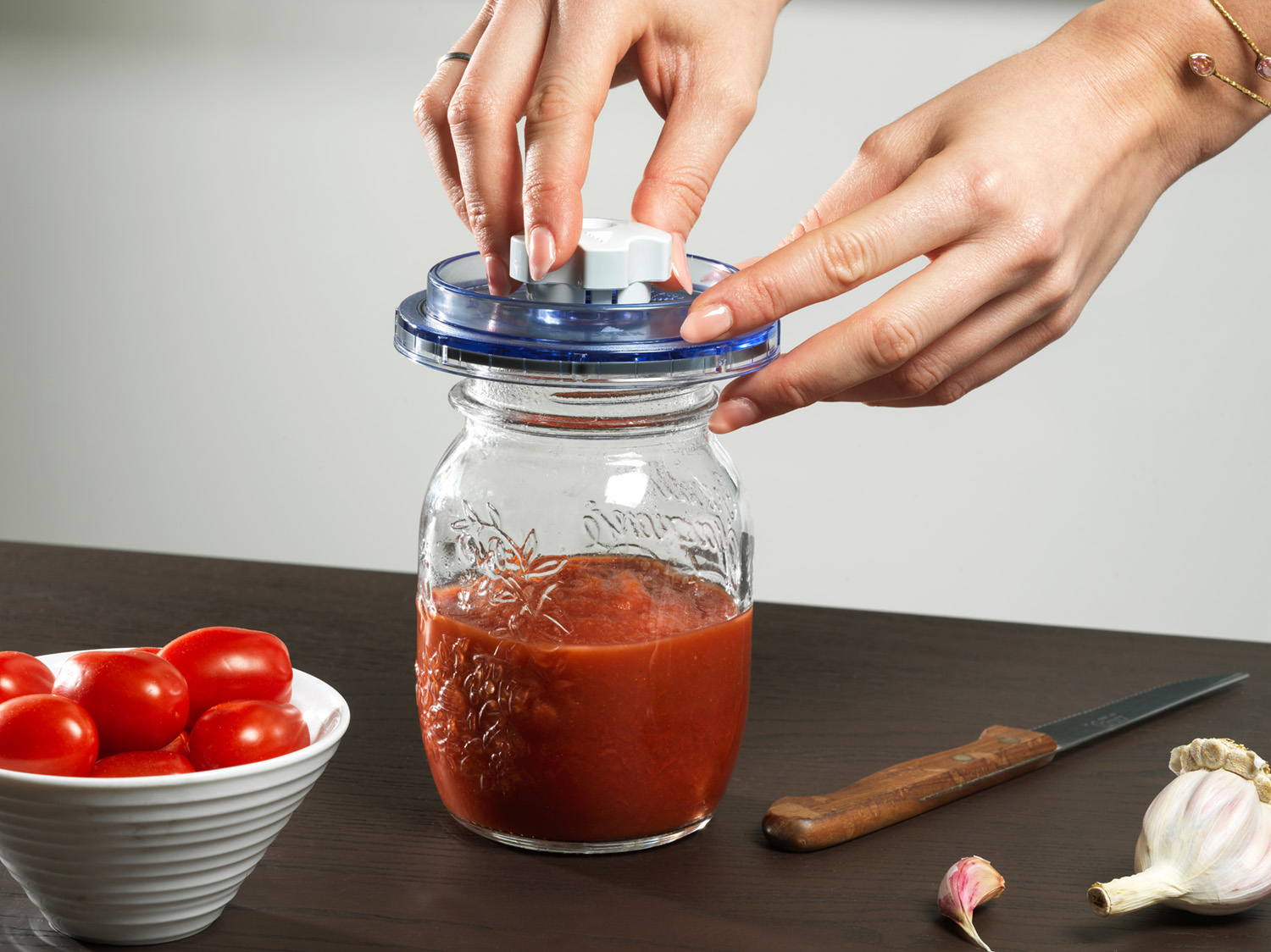 Mani che sigillano un barattolo di salsa di pomodoro. Pomodori, aglio e un coltello sono sul tavolo. Il barattolo è trasparente con coperchio blu.