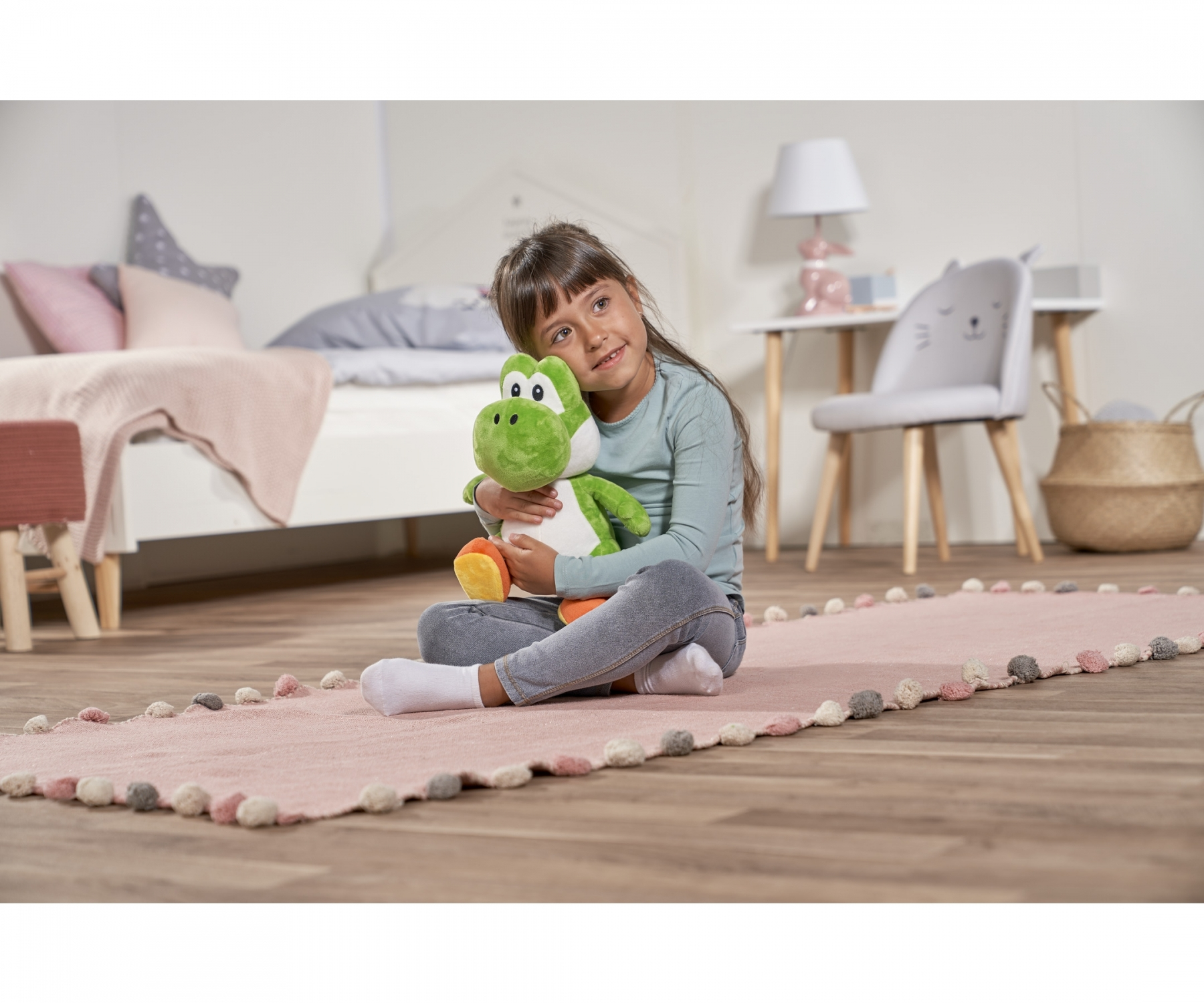 Une fille serre un jouet en peluche Yoshi en s'asseyant sur un tapis rose. Un lit et une chaise sont en arrière-plan.