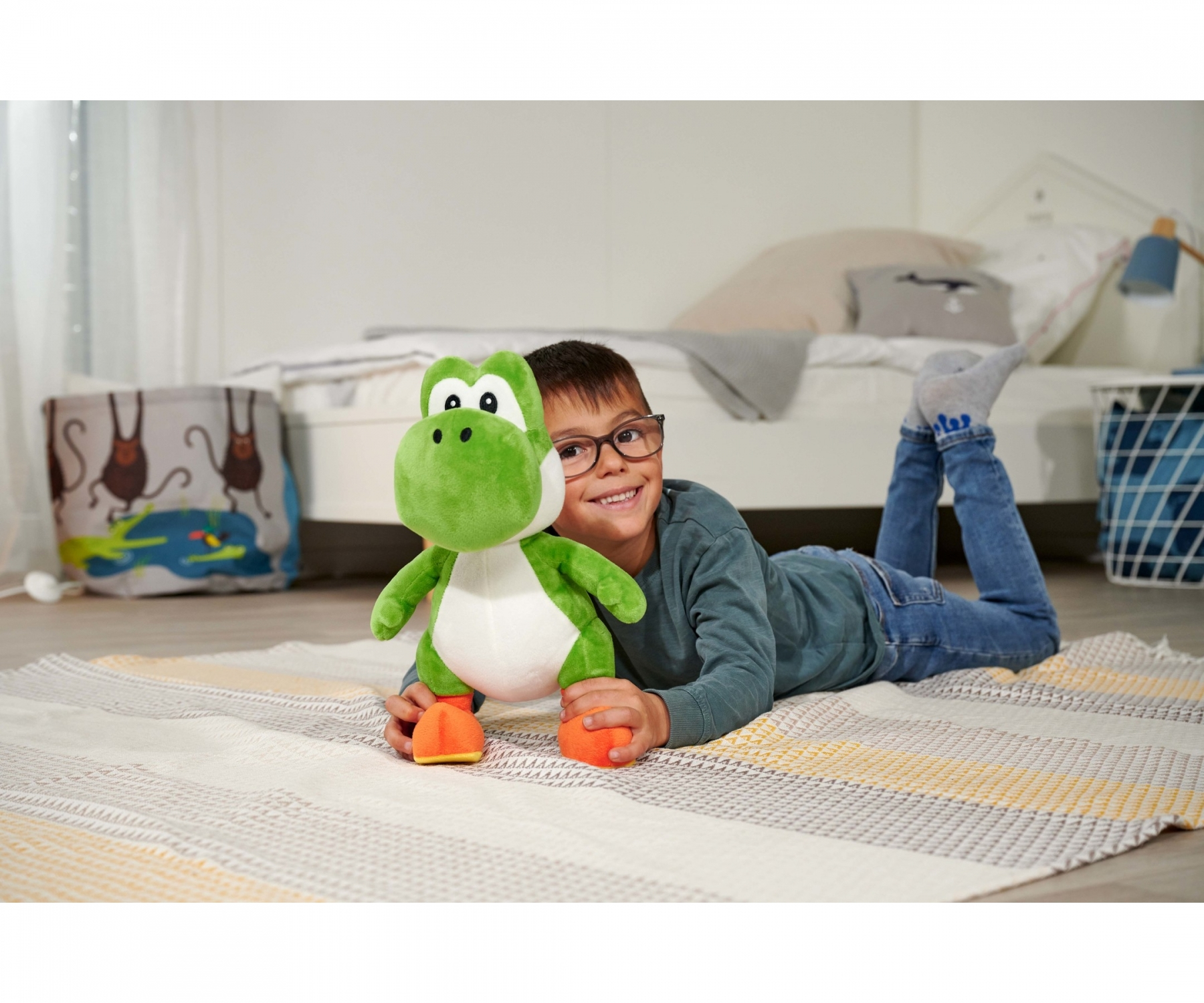 Un garçon avec des lunettes sourit, tenant une peluche Yoshi. Il est allongé sur un tapis, avec un lit et une décoration de chambre en arrière-plan.