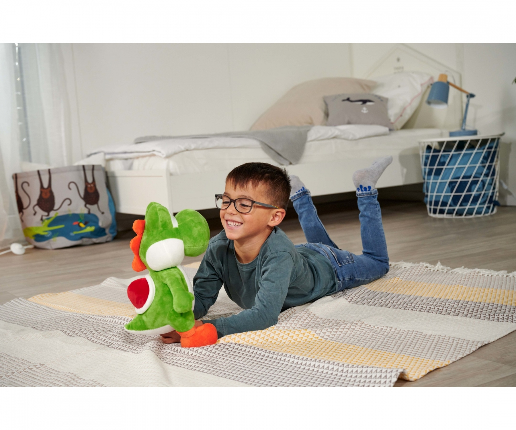 Un garçon avec des lunettes sourit en tenant une peluche Yoshi. Il est allongé sur un tapis, un lit et une décoration de chambre en arrière-plan.