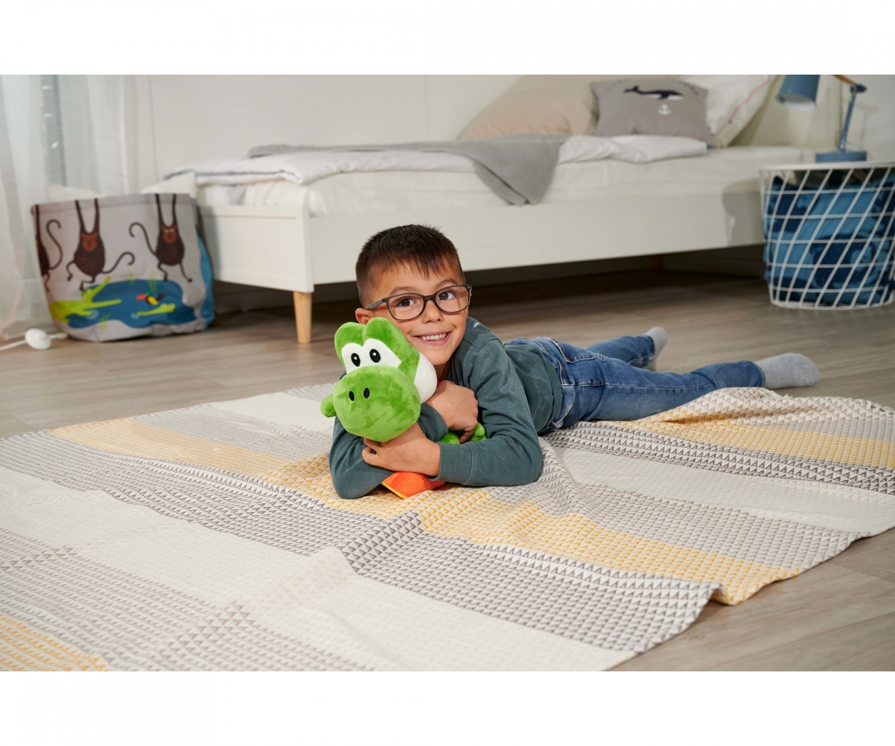 Un garçon avec des lunettes est allongé sur un tapis, embrassant une peluche Yoshi verte. Un lit et un panier sont en arrière-plan.