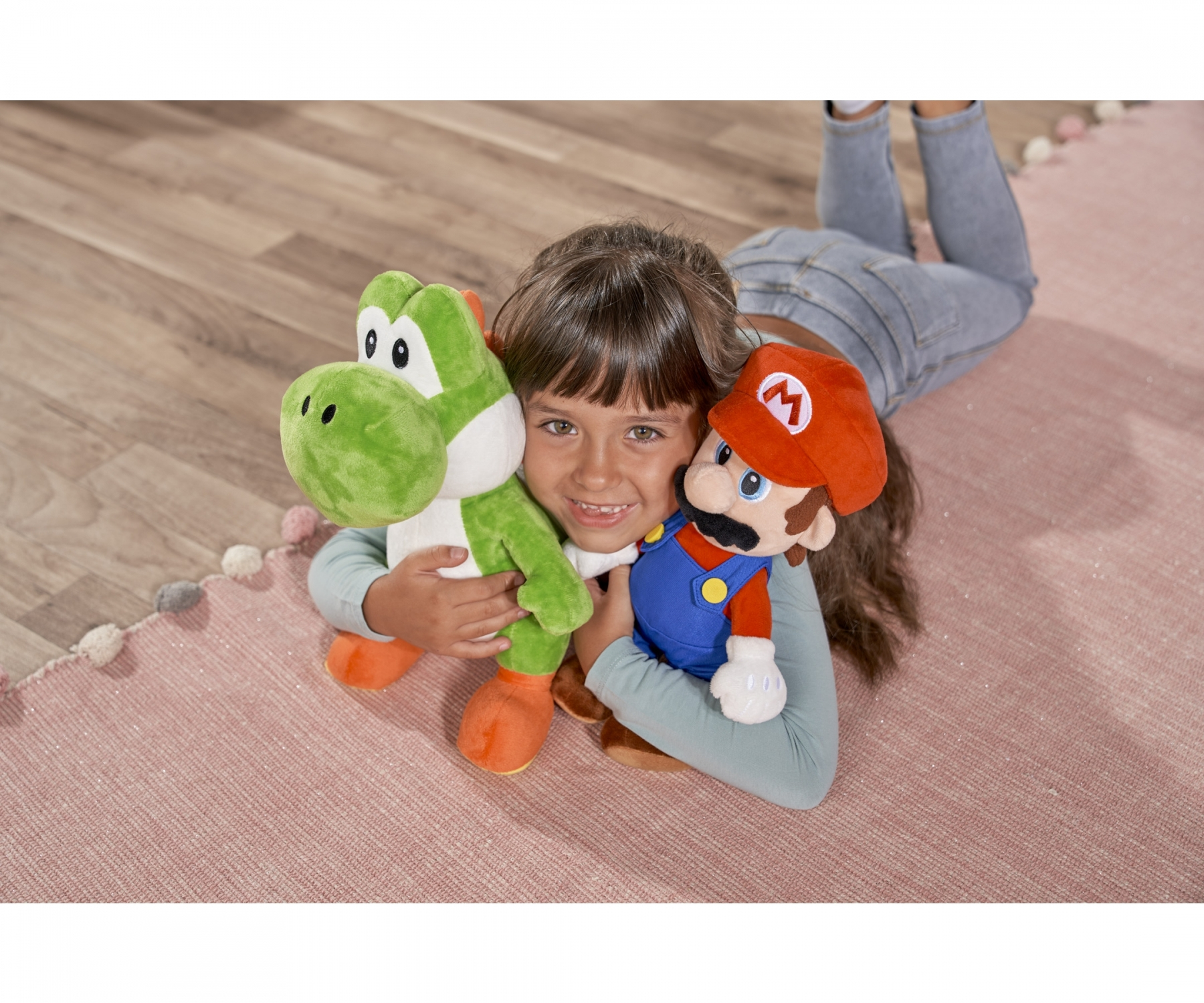 Une fille sourit en tenant des peluches Yoshi et Mario. Elle est allongée sur un tapis rose avec une bordure en pompons. Plancher en bois en arrière-plan.