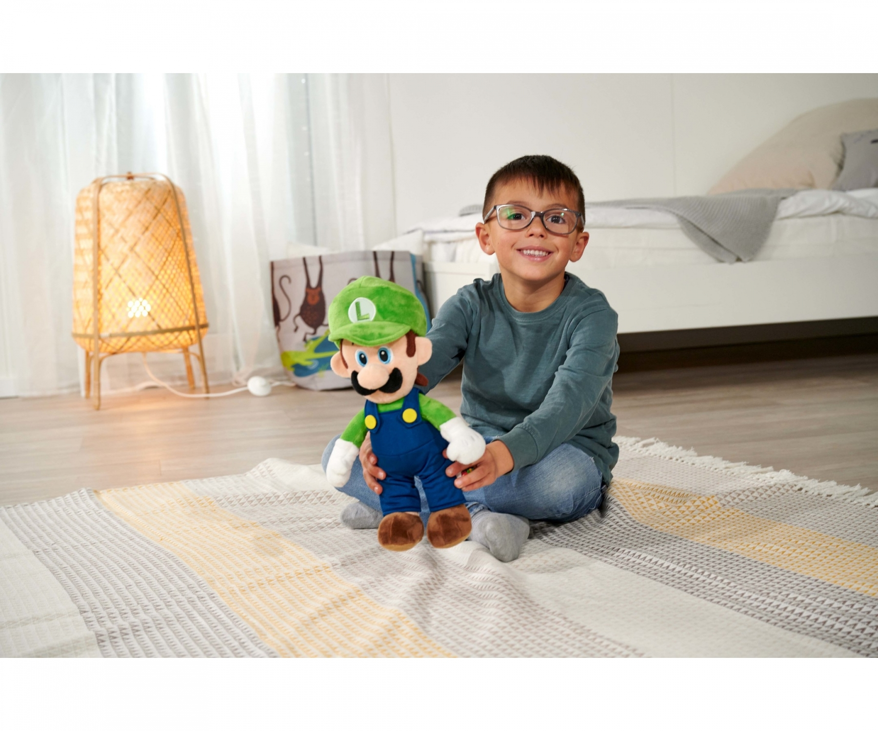 Ein Junge mit Brille lächelt und hält einen Luigi-Plüsch. Er sitzt auf einem gestreiften Teppich. Lampe und Bett im Hintergrund.