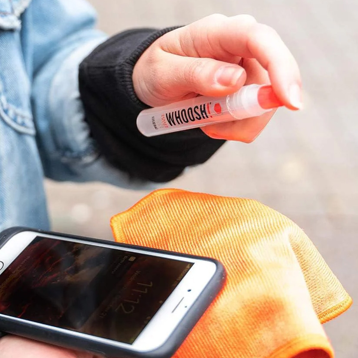Eine Hand hält eine Sprühflasche über einem Telefon und einem orangefarbenen Tuch. Der Telefonbildschirm zeigt 11:12. Die Flasche sagt WHOOSH!.