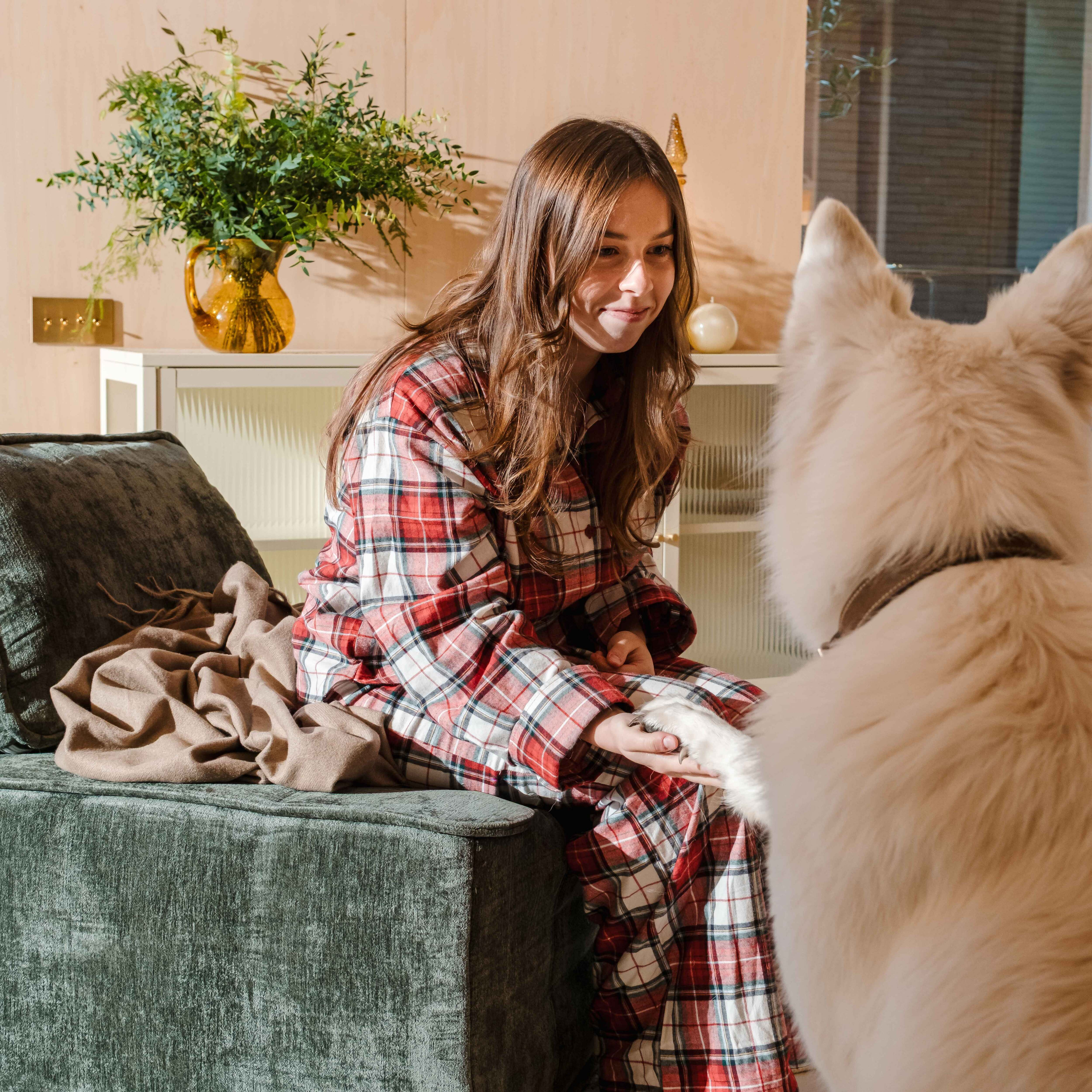 Eine Frau im Pyjama sitzt und berührt die Pfote eines weißen Hundes. Ein Vasen mit Pflanzen und eine Couch im Hintergrund.