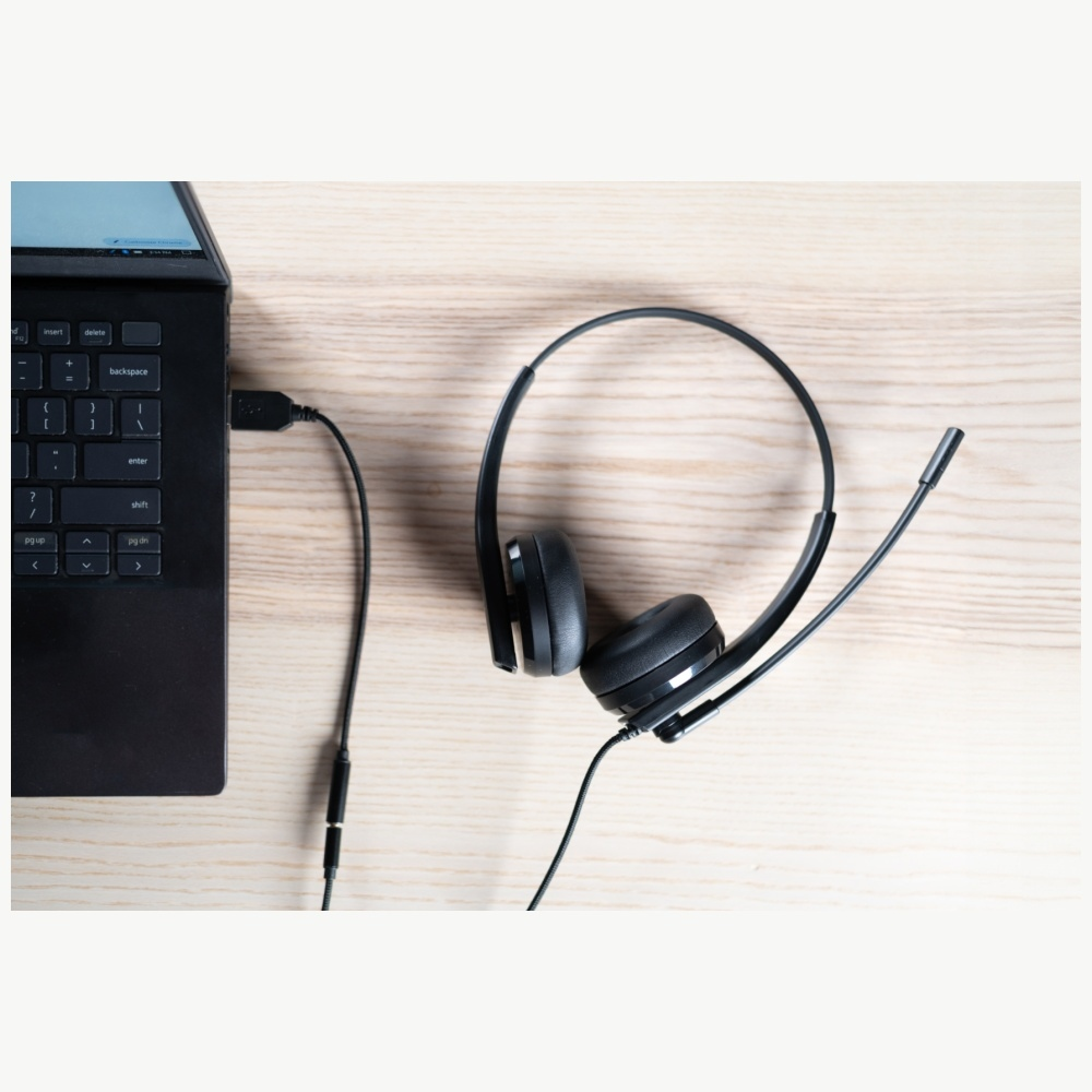 Ein schwarzer Laptop mit Headset liegt auf einer Holzoberfläche. Das Headset hat ein Mikrofon und ist mit dem Laptop verbunden.