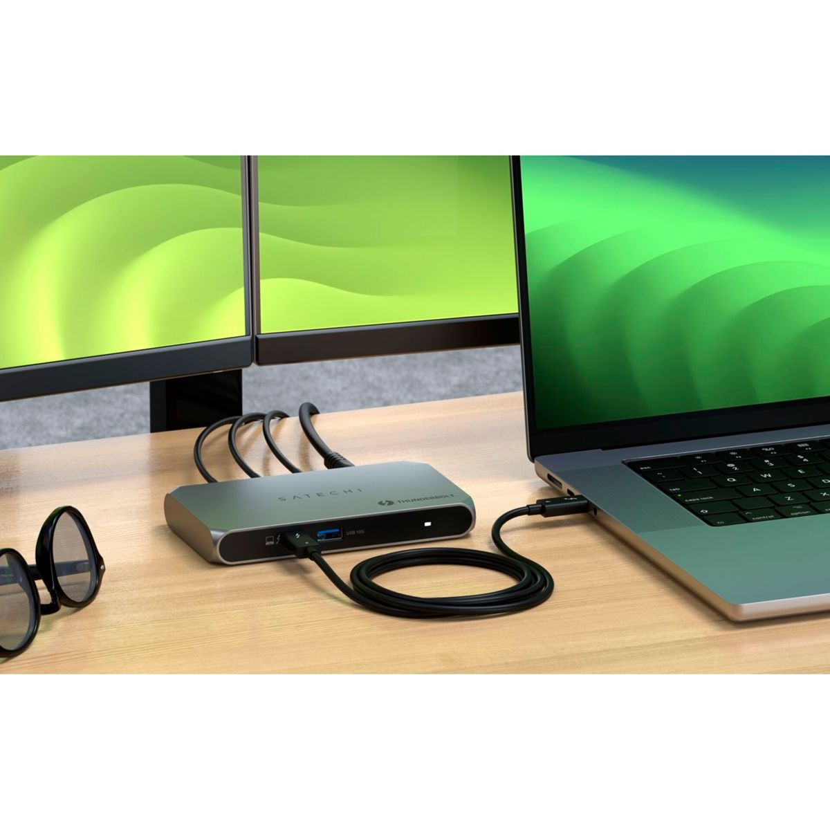 Un laptop collegato a una docking station con più monitor. Occhiali sulla scrivania. Tutto su una scrivania di legno con sfondo verde.