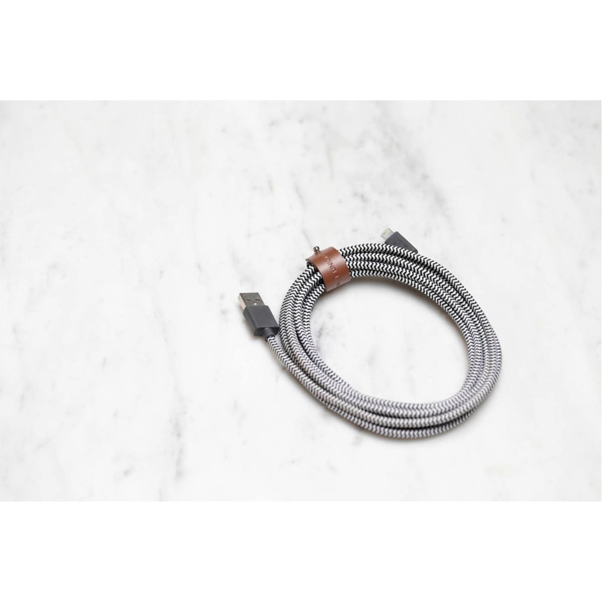 Ein geflochtenes USB-Kabel in Grau und Weiß, auf einer weißen Marmoroberfläche aufgerollt. Es hat ein braunes Lederband und schwarze Anschlüsse.
