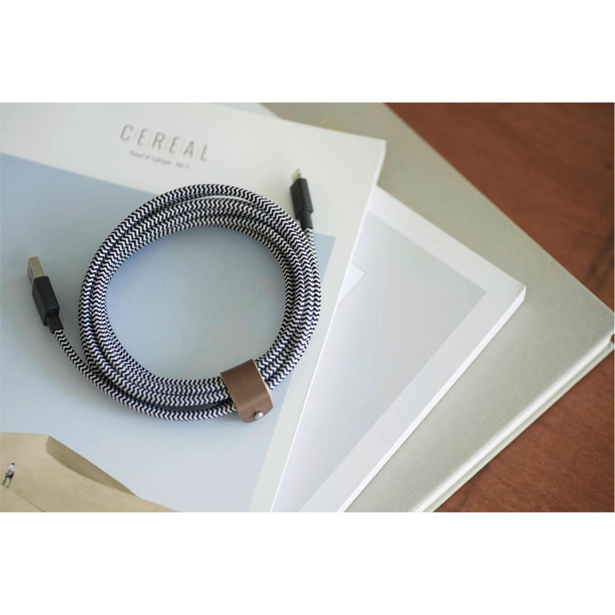 Ein gewendeltes schwarz-weißes USB-Kabel mit Lederriemen liegt auf Büchern. Eine Zeitschrift mit dem Wort CEREAL ist sichtbar.