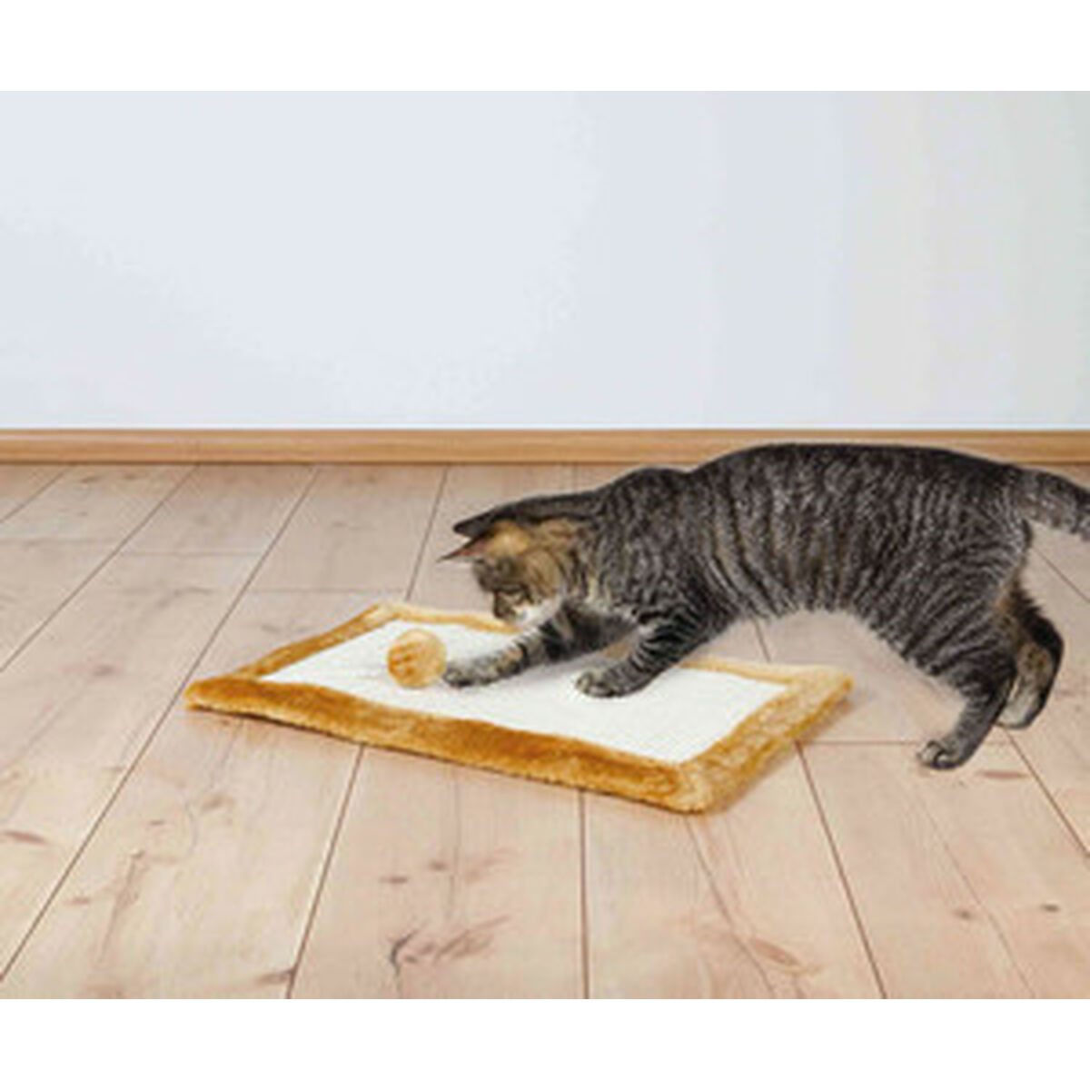 Un gatto tigrato gioca con un giocattolo su un tappetino. Il tappetino è su un pavimento in legno e lo sfondo è un muro bianco.