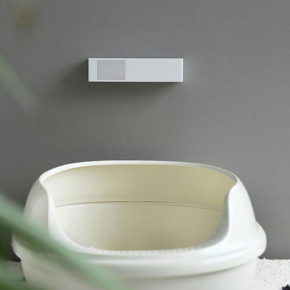 Biały prostokątny obiekt z otworami na szarej ścianie. Poniżej widoczny biały żwirek dla kota.